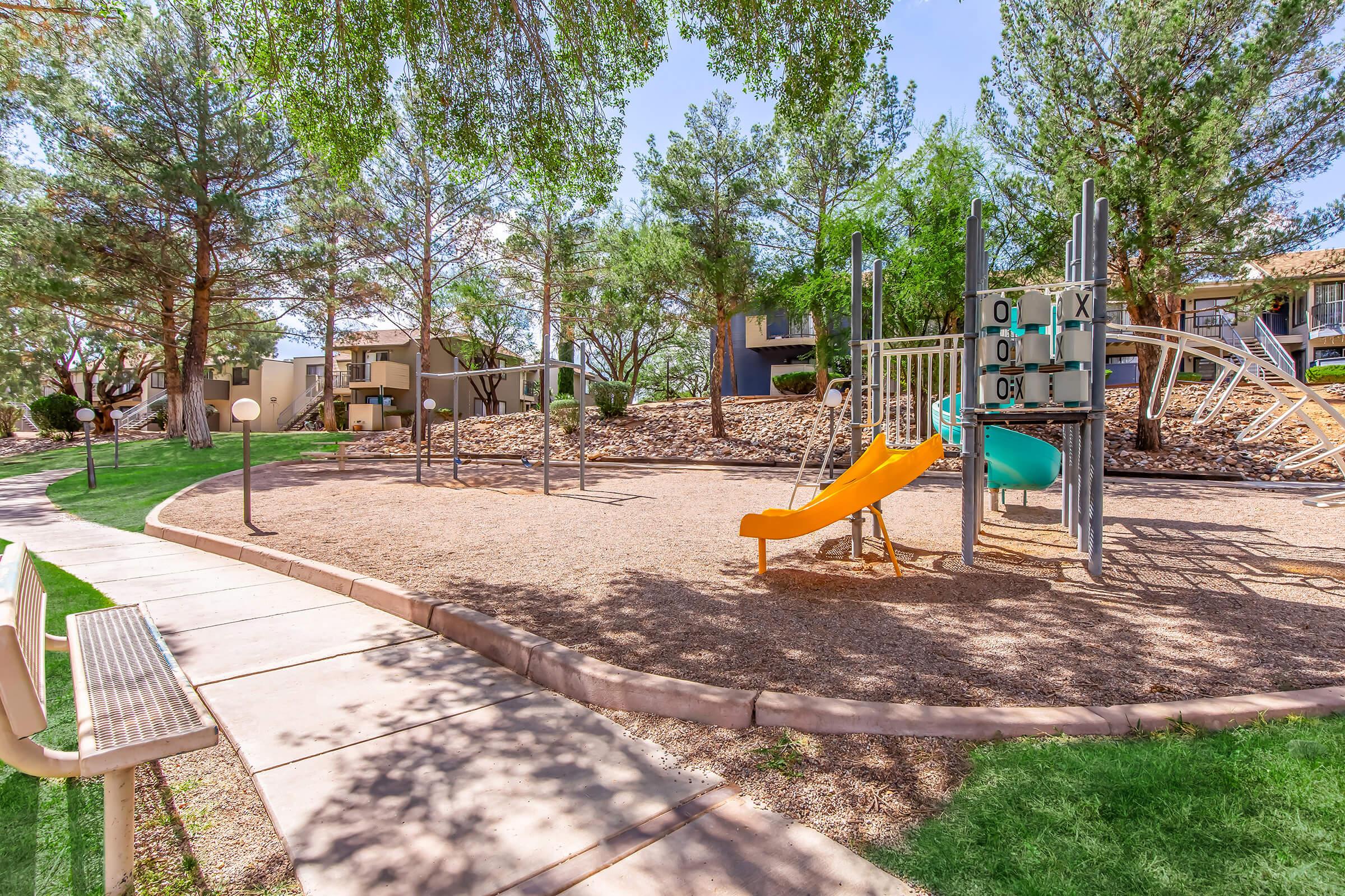 A sunny playground surrounded by trees, featuring a yellow slide, a blue slide, and climbing structures. The area is sandy with a walking path and benches nearby, providing a pleasant outdoor space for children to play.
