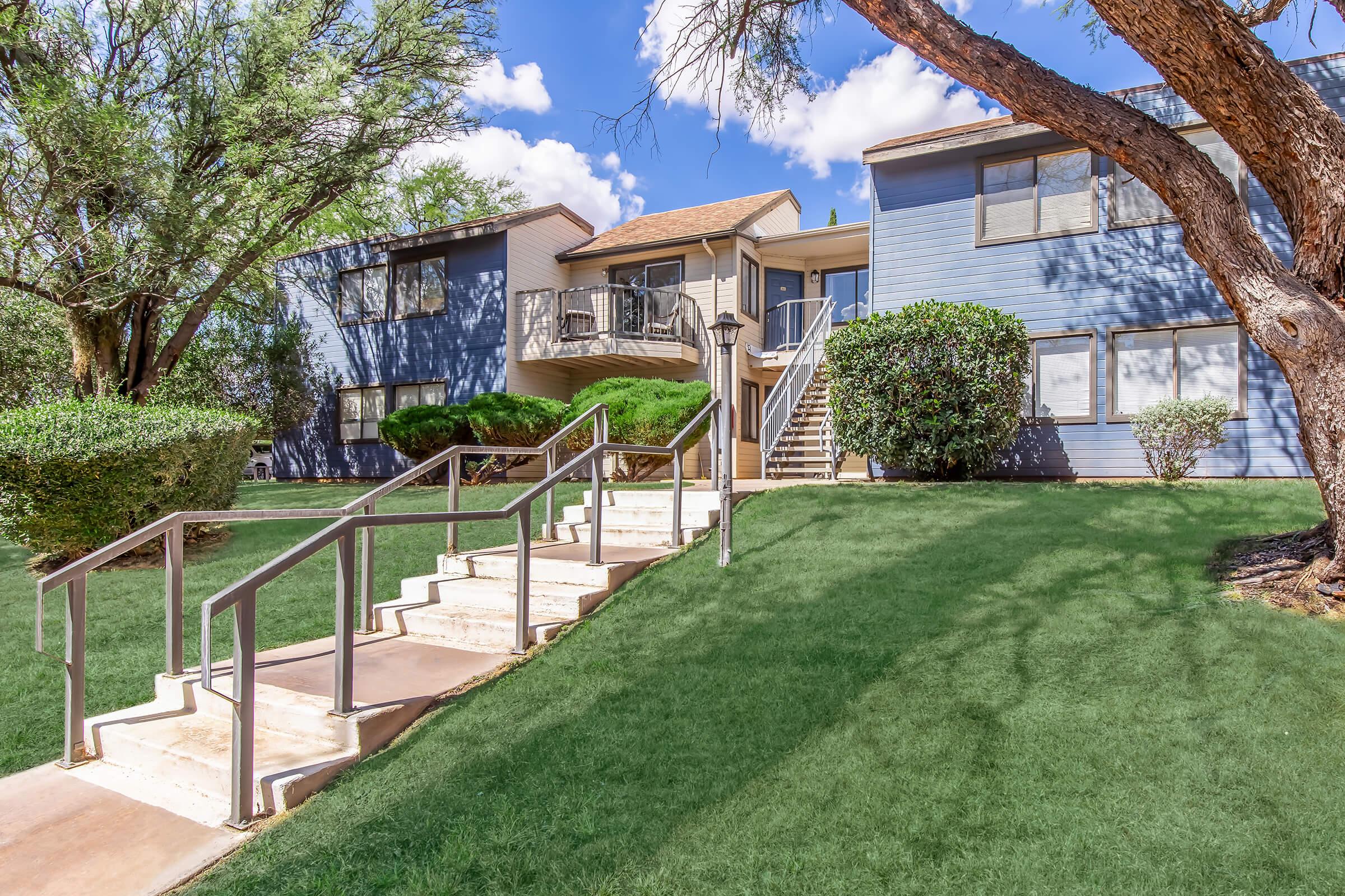 A well-maintained apartment complex with two-story buildings surrounded by lush greenery. There are stairs leading up to the entrance, flanked by neatly trimmed bushes. The sky is clear with a few fluffy clouds, and a lamp post stands near the path, adding to the inviting atmosphere.