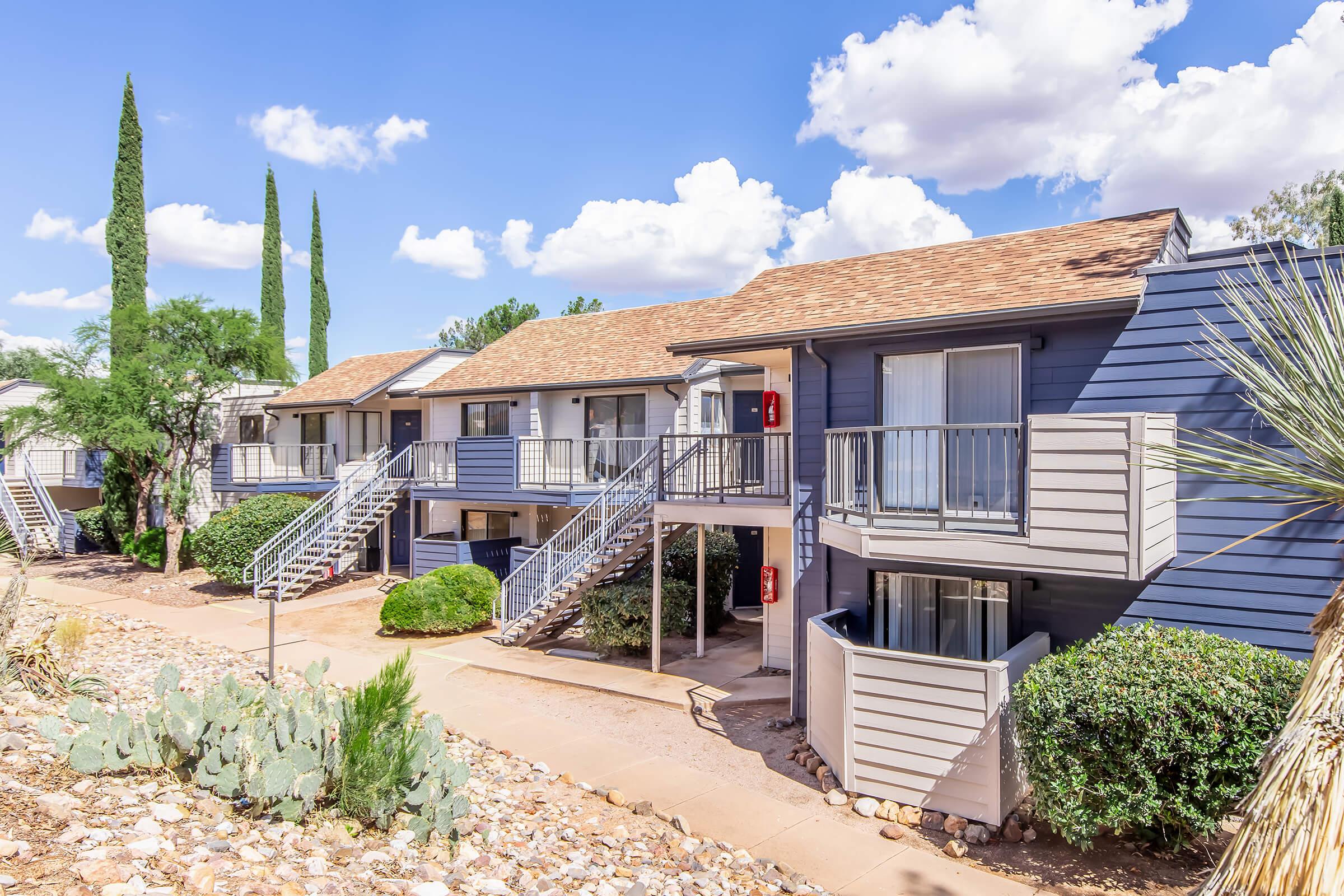 A view of a two-story apartment complex with a row of units featuring balconies. Several tall, narrow cypress trees are visible alongside the building, and desert landscaping with rocks and cacti is present in the foreground. The sky is partly cloudy.