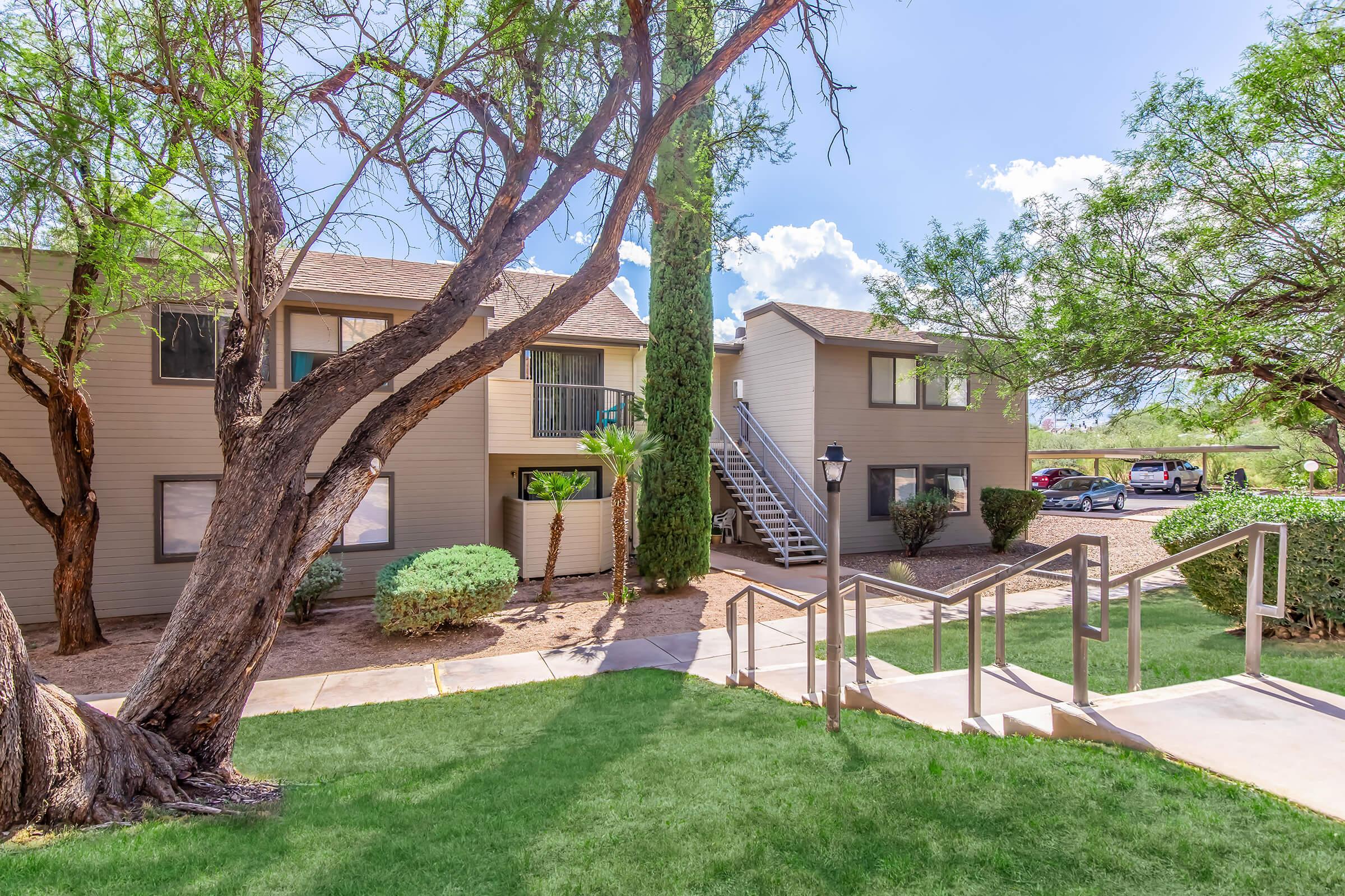 A view of a landscaped apartment complex featuring two buildings, one with a staircase leading up, outlined by grass and shrubs. The area is shaded by mature trees under a partly cloudy sky, creating a pleasant outdoor environment.