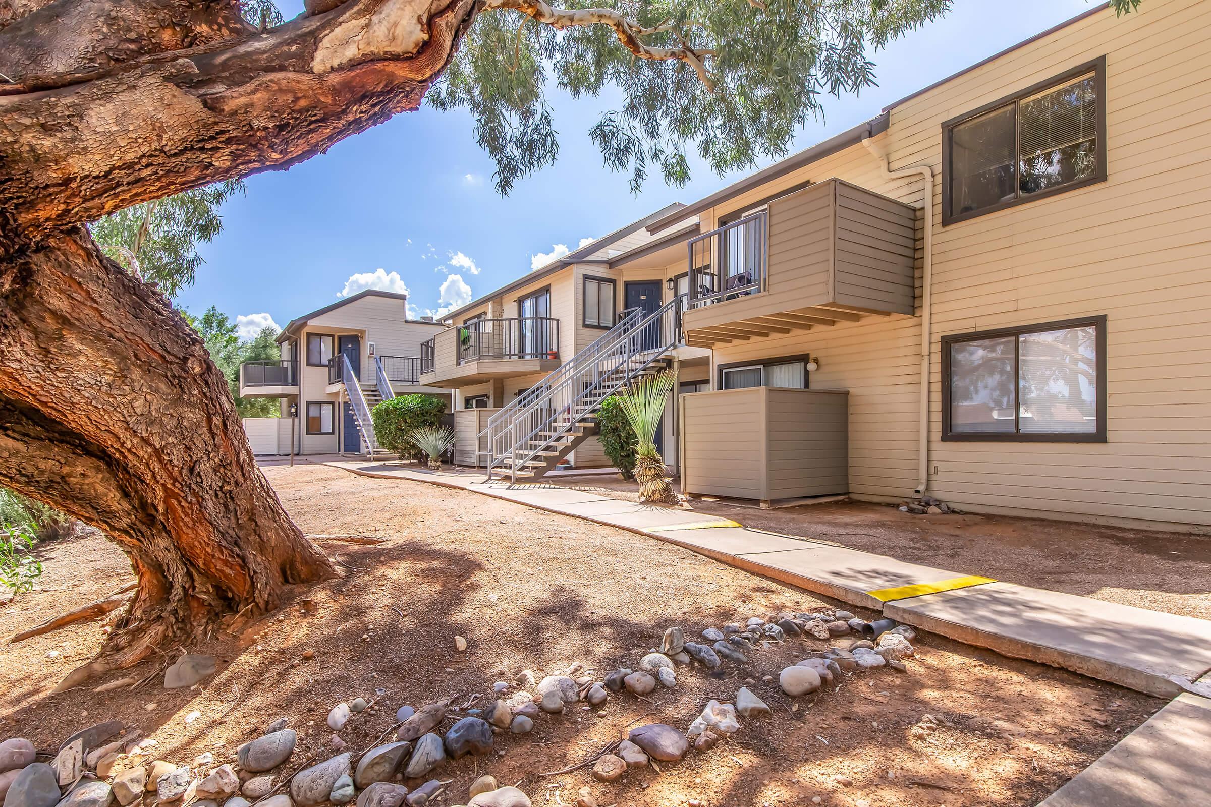 A view of a residential apartment complex featuring two-story buildings with balconies. A large tree and landscaped area with rocks are in the foreground, alongside a pathway leading to the entrance of the apartments. The sky is bright with scattered clouds.