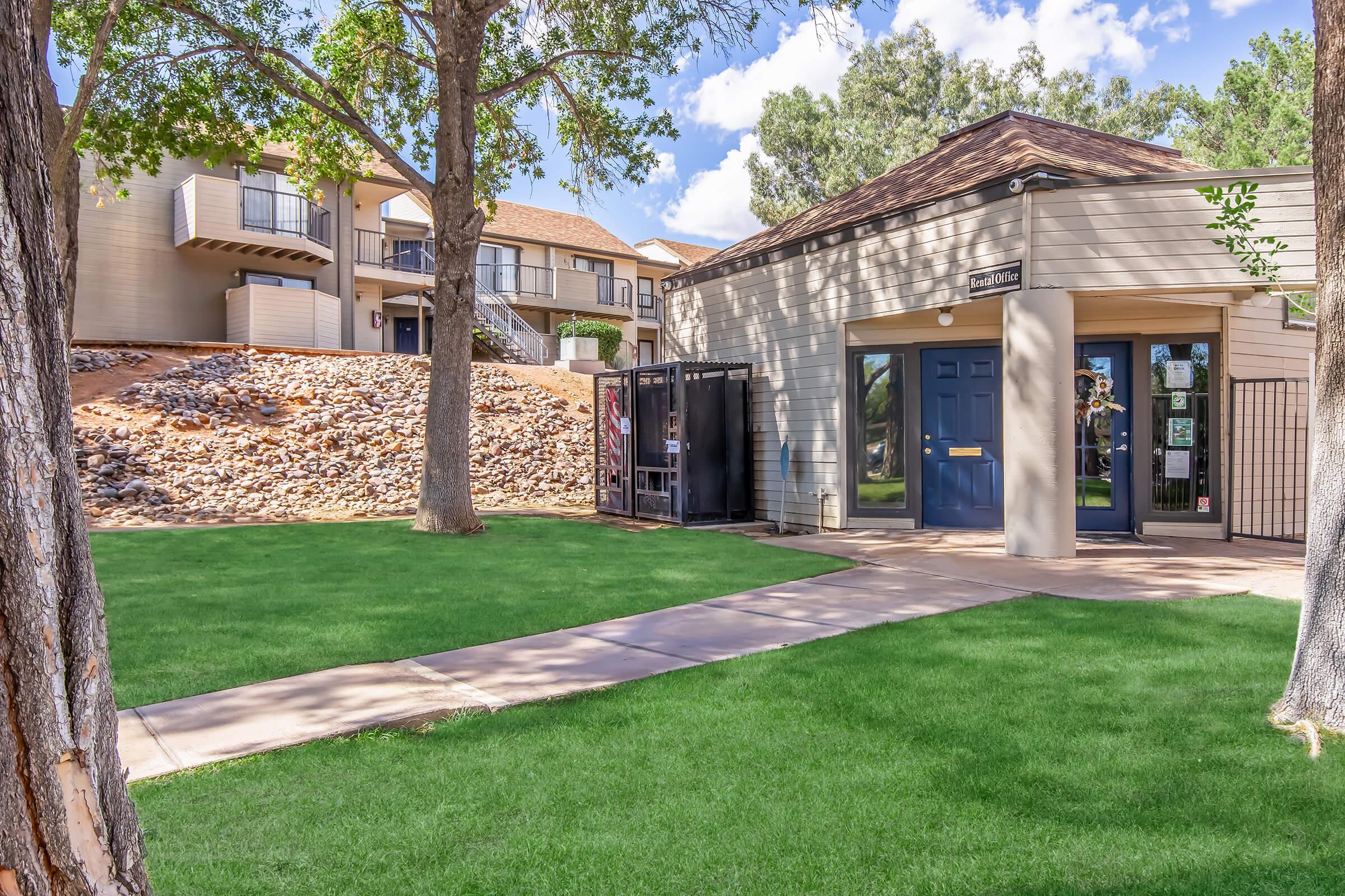A well-maintained outdoor area featuring green grass, a paved walkway, and a building with a blue door. In the background, there are additional structures with balconies, surrounded by trees and a rocky hillside. The scene has a clear blue sky with fluffy clouds.