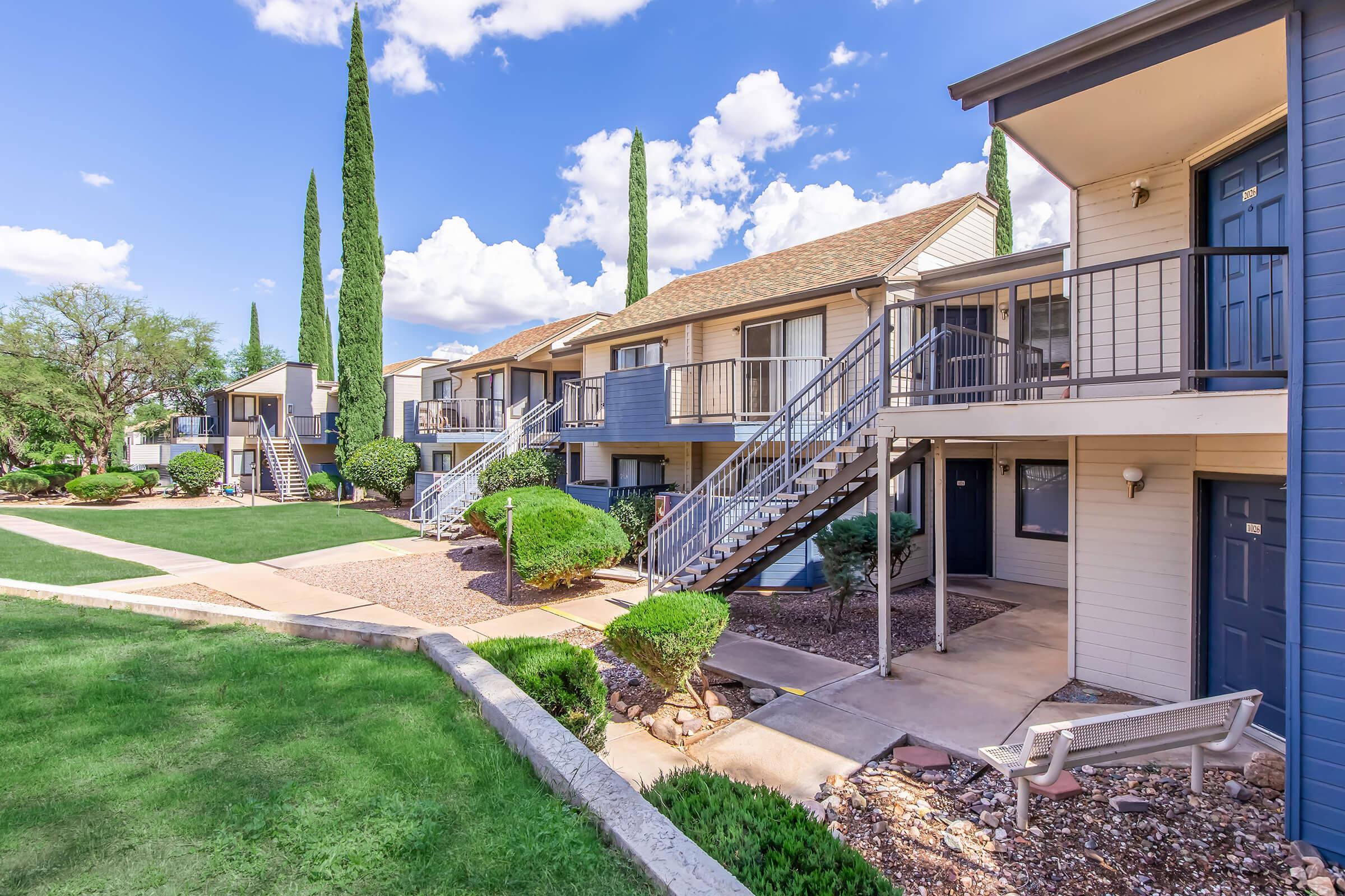 A view of two-story apartment buildings surrounded by well-maintained landscaping, featuring green lawns, shrubs, and tall cypress trees under a bright blue sky with fluffy clouds. The buildings have balconies and stairs leading to the entrances.
