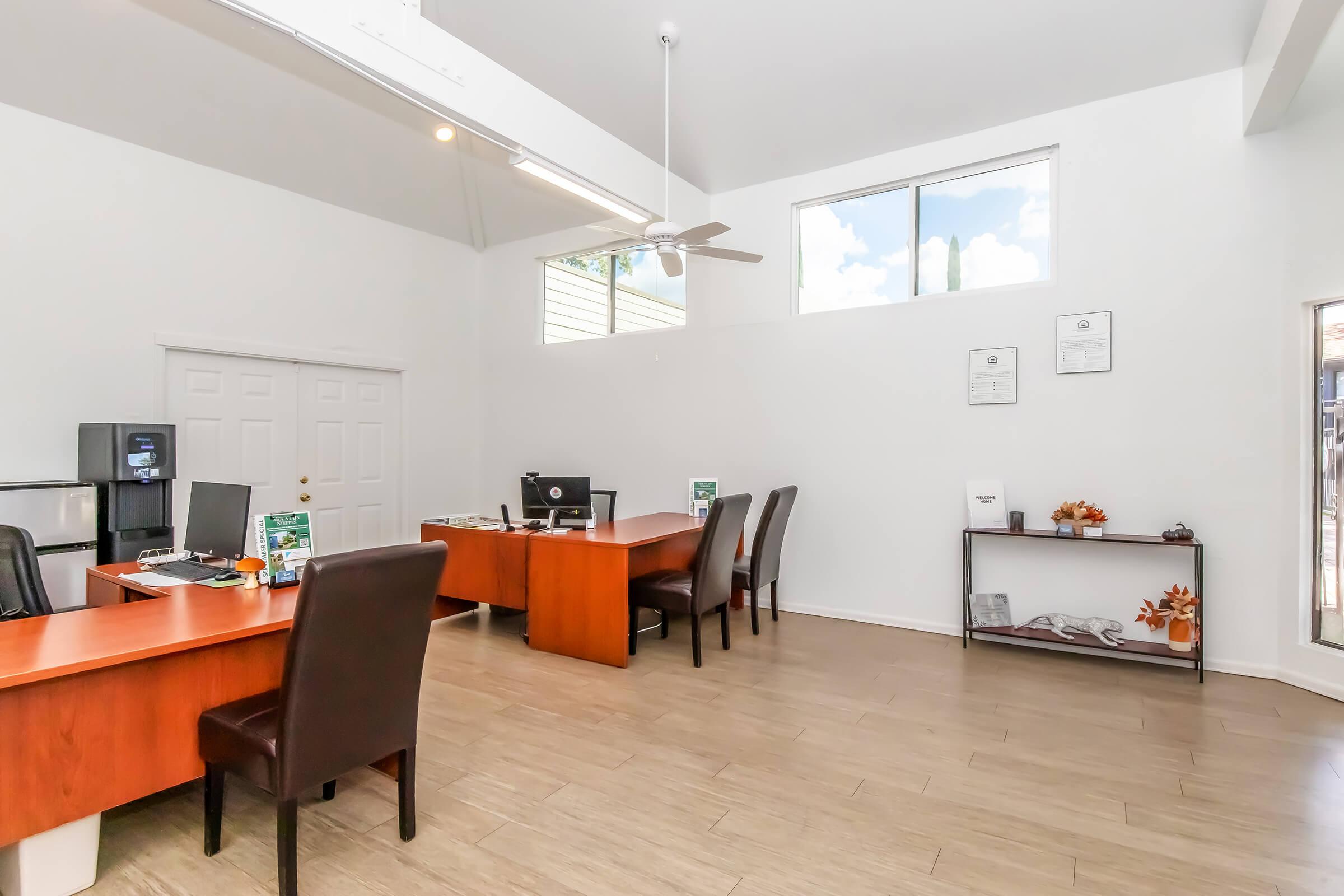 Interior view of a modern office space featuring two wooden desks with chairs, a computer setup, and a reception area. The room has large windows allowing natural light, a ceiling fan, and minimal decorations on a side shelf. The walls are painted white, creating a spacious and bright atmosphere.