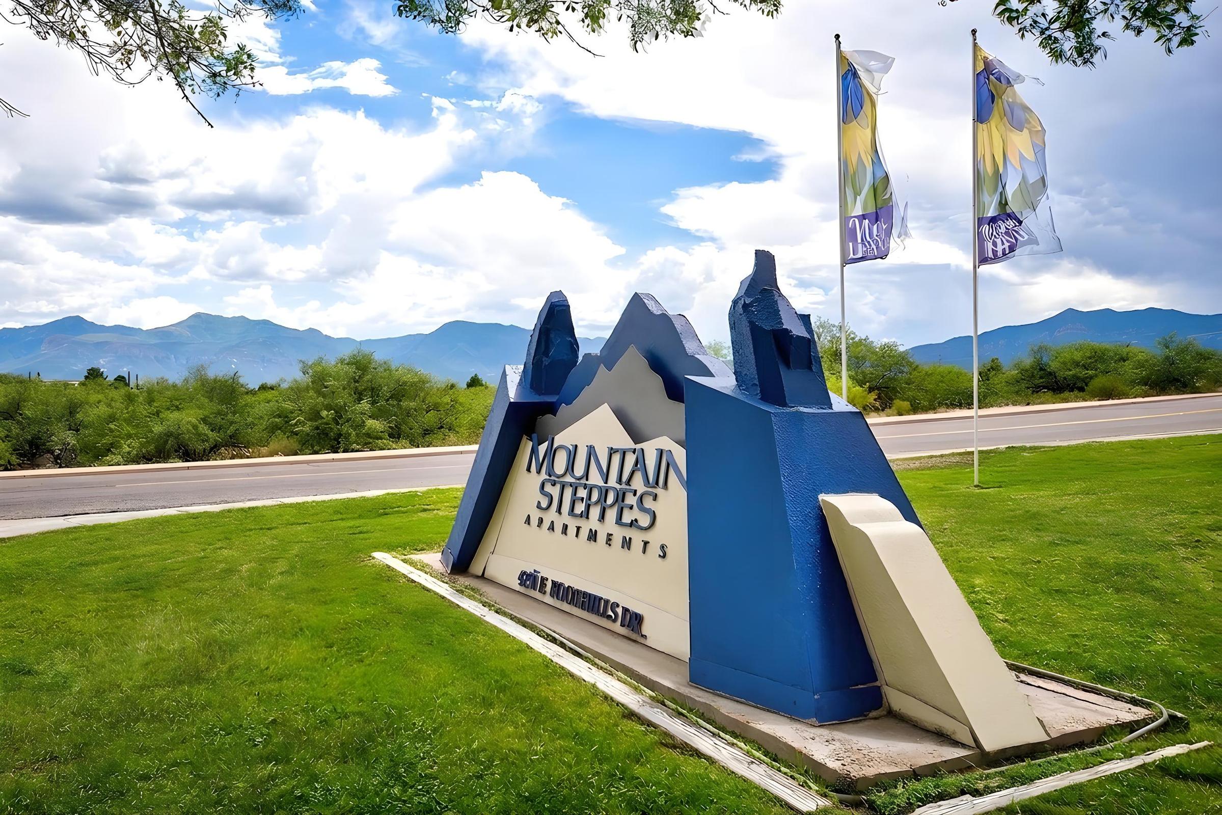 Sign for Mountain Steppes Apartments, featuring a stylized mountain design in blue and white. There are two flags next to the sign, with a background of green grass and mountains under a partly cloudy sky. The address displayed is 926 Mountain View St.
