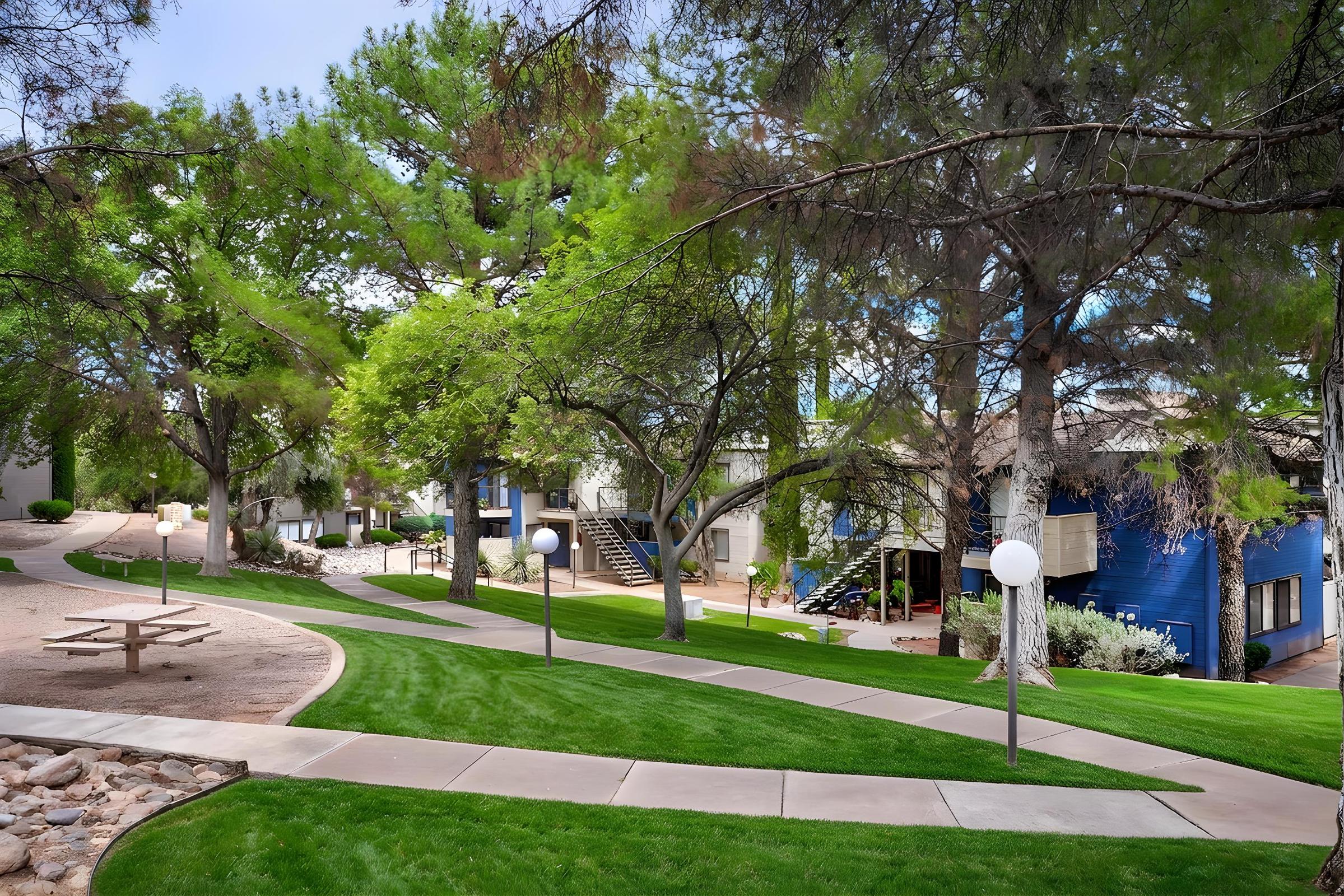 A landscaped pathway winds through a residential area with green lawns and tall trees. There are several apartment buildings visible, featuring a mix of colors, including blue. White lamp posts line the path, and a picnic table is positioned nearby, creating a serene outdoor environment.