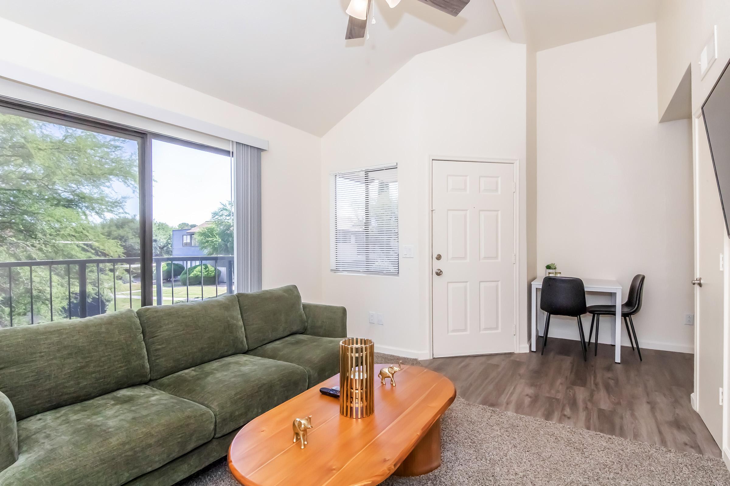 A cozy living room featuring a green sofa, a wooden coffee table, and a small workspace with a chair and desk. Large windows allow natural light to fill the space, with a view of greenery outside. The room has a high ceiling and modern decor, creating a welcoming atmosphere.