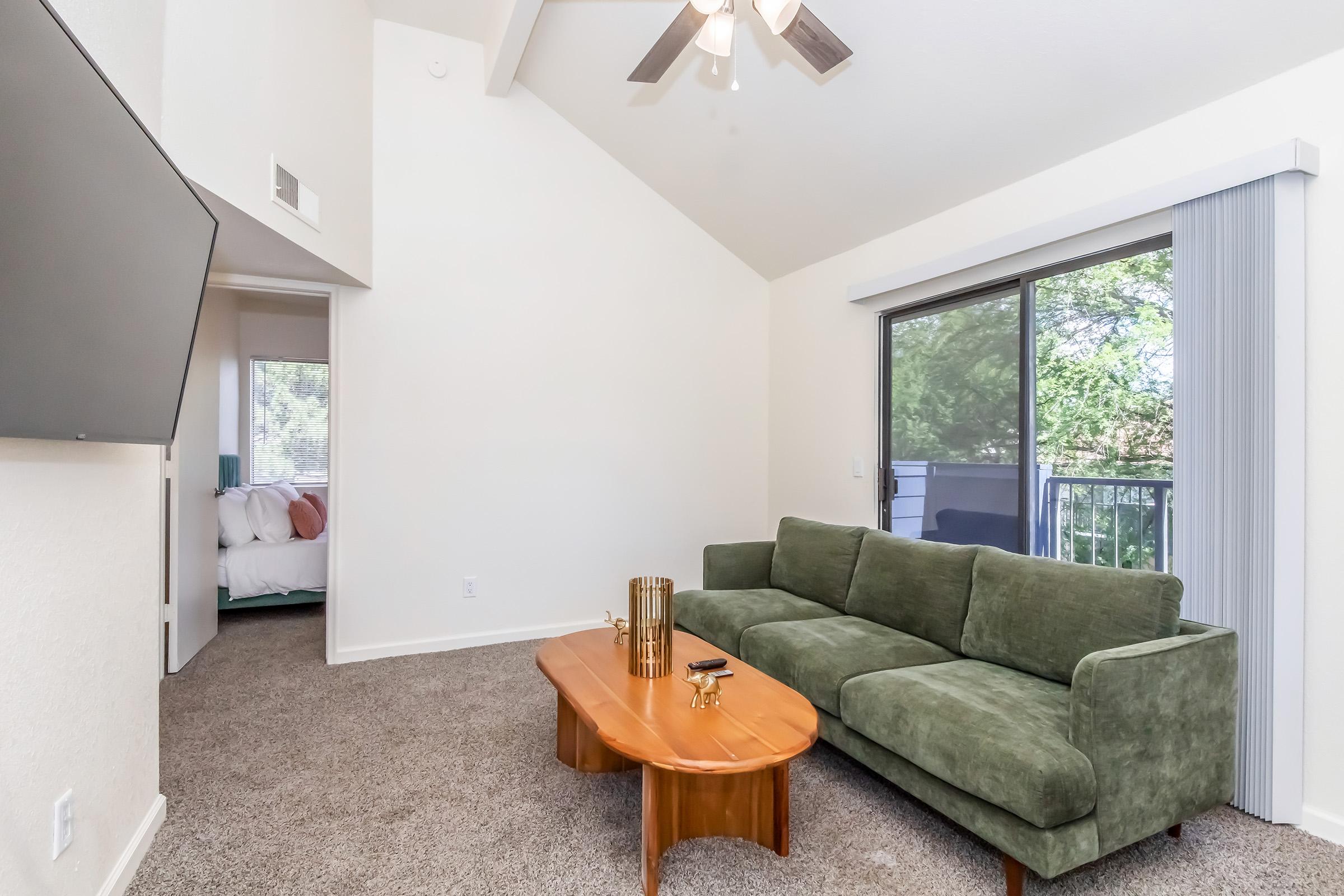 Living room featuring a green couch and a wooden coffee table, set against a backdrop of light-colored walls and a ceiling fan. A large window allows natural light in, offering a view of greenery outside. In the background, a doorway leads to a bedroom with a bed visible. The flooring is carpeted.