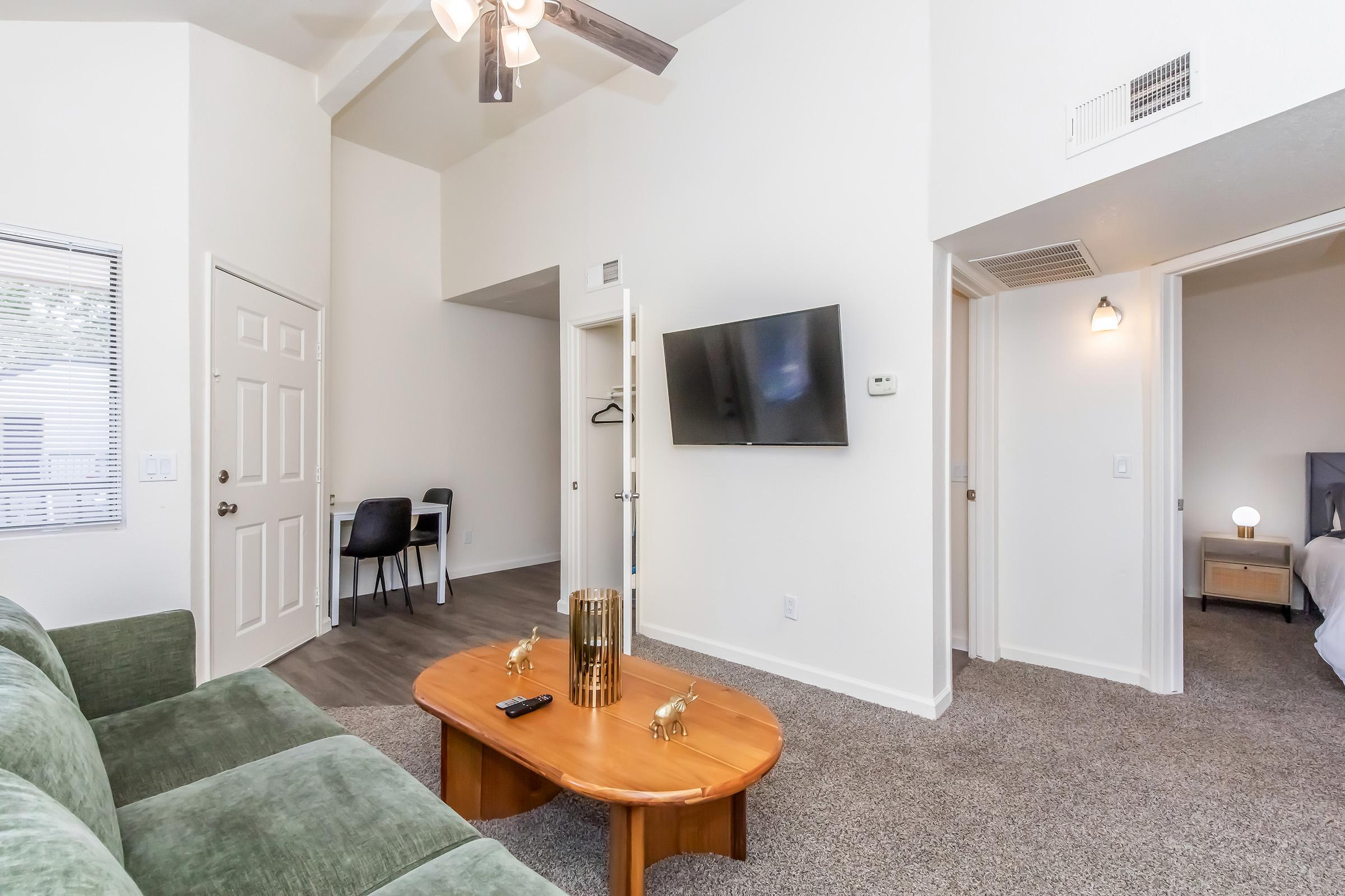 A cozy living room featuring a green sofa, a wooden coffee table, and a flat-screen TV mounted on the wall. The room has a ceiling fan and a light-colored interior. An open doorway leads to another room, while a chair is positioned nearby. The carpet is light gray, creating a warm atmosphere.