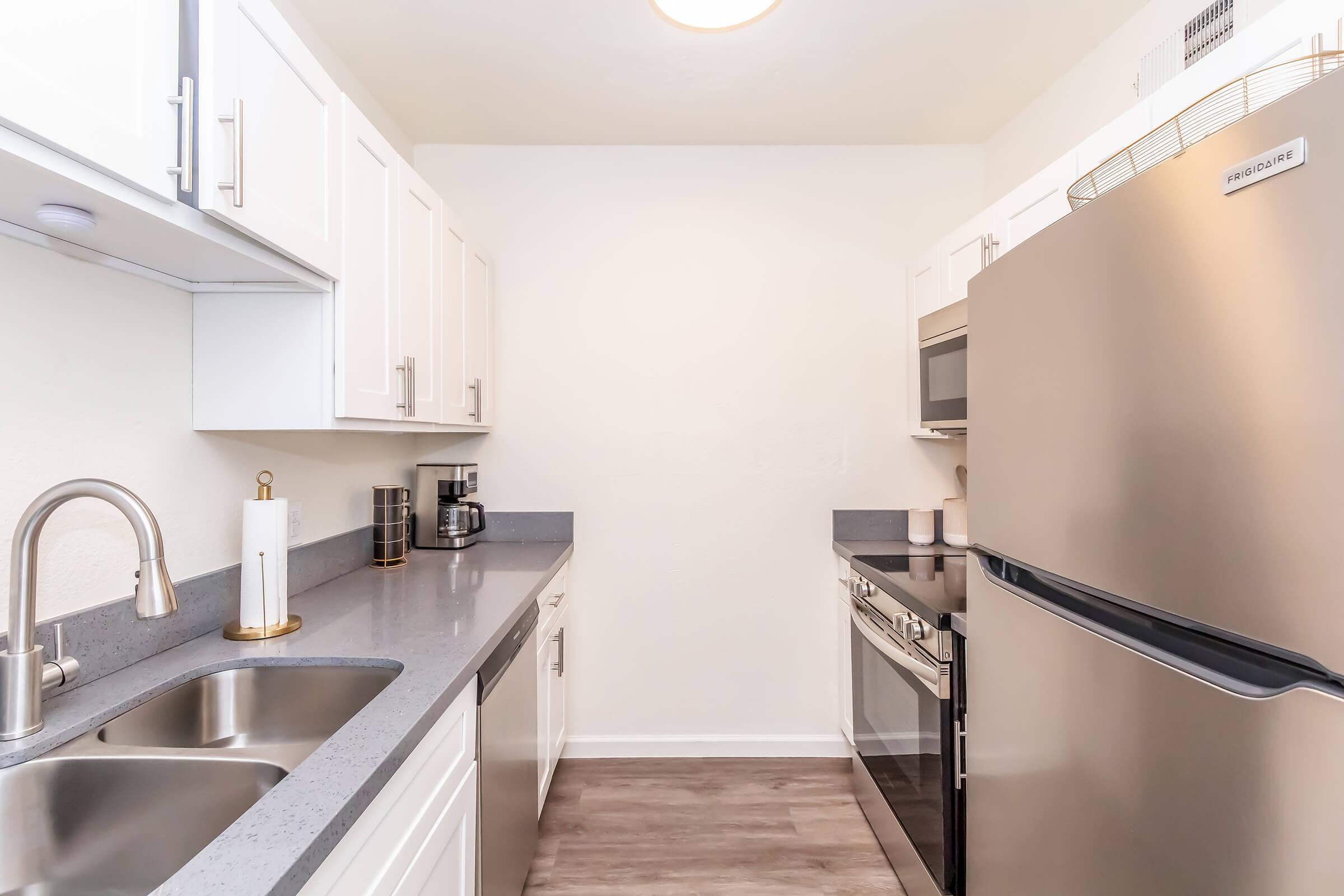 Modern kitchen featuring stainless steel appliances including a refrigerator and microwave, gray countertops, white cabinets, and a coffee maker. The layout is minimalist with a clean aesthetic, showcasing a bright, uncluttered space suitable for cooking and dining.