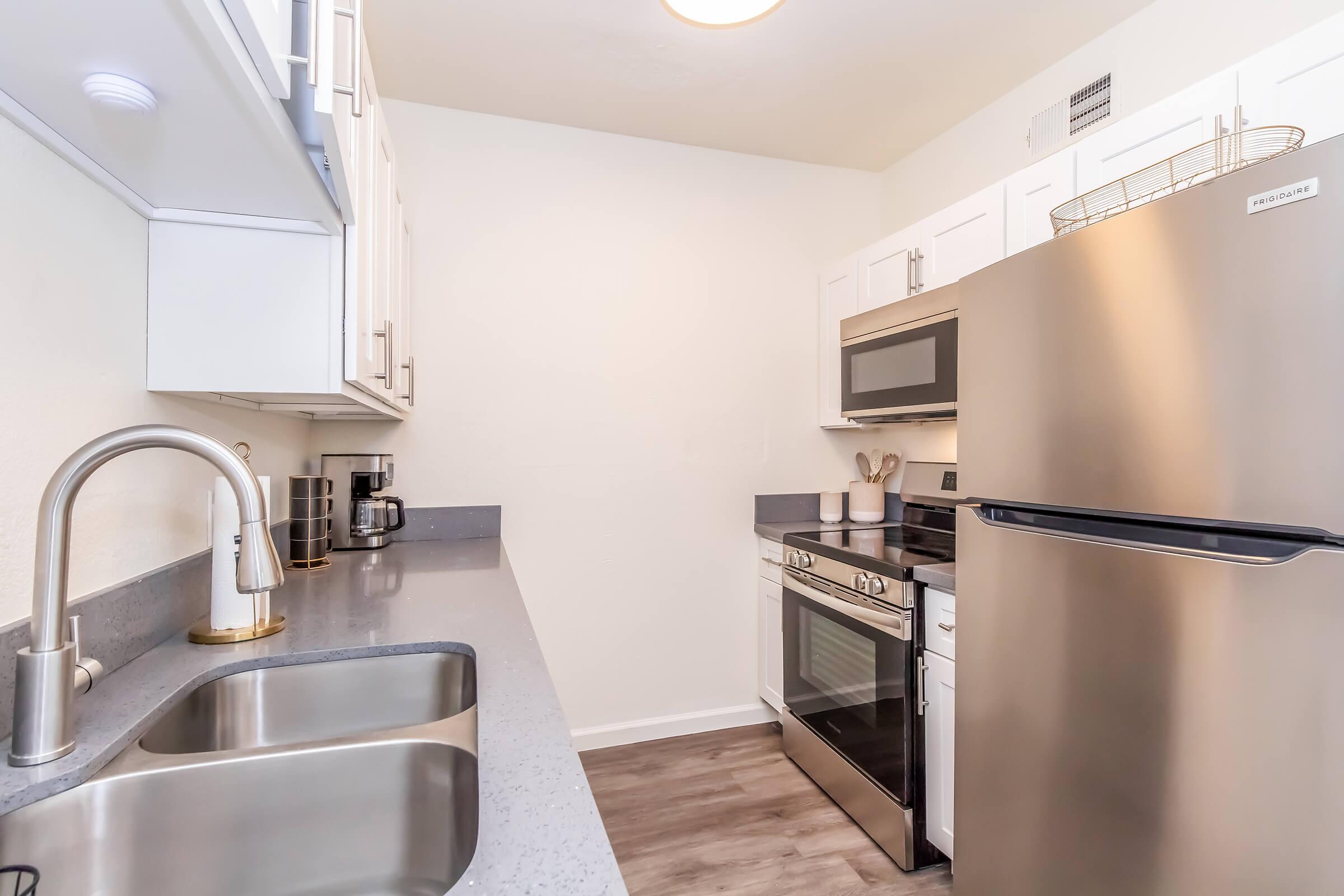 A modern kitchen featuring stainless steel appliances, including a microwave, oven, and refrigerator. The design includes a double sink with a sleek faucet, gray countertops, and white cabinetry. There’s a coffee maker and wooden utensils arranged neatly, creating a functional and stylish space.