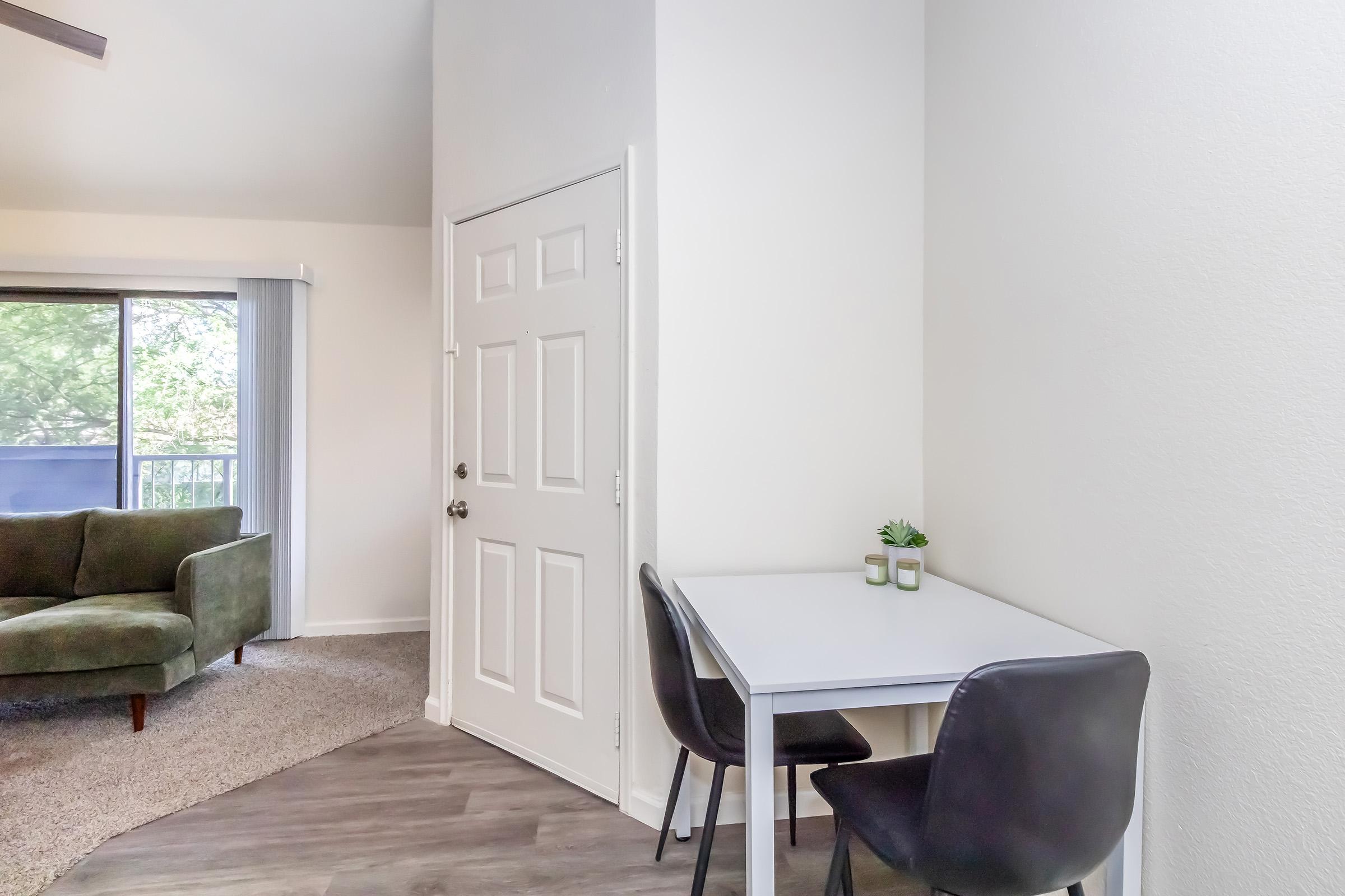 A cozy corner of an apartment featuring a small white table with two black chairs, a decorative plant on the table, and a doorway leading outside. Natural light floods in from a sliding glass door, illuminating the space which includes a green sofa and light-colored walls.