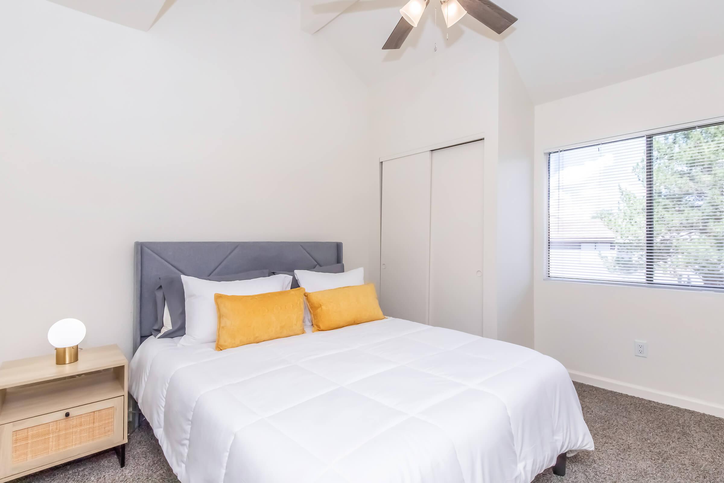A well-lit bedroom featuring a queen-size bed with white linens and yellow accent pillows. There is a bedside table with a lamp and a closet in the background. A window reveals natural light, enhancing the warm and inviting atmosphere of the room.