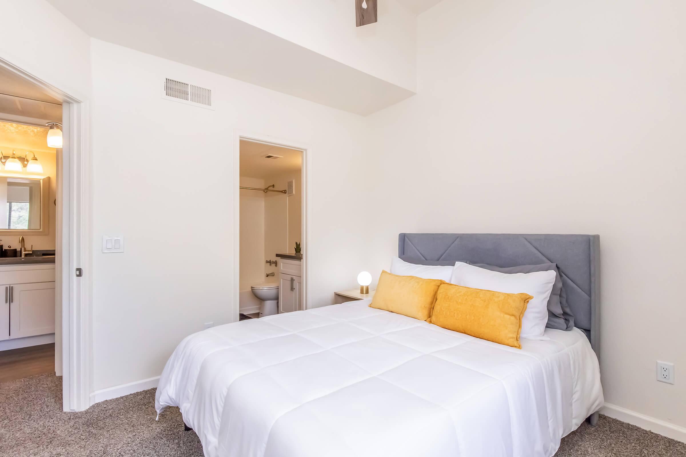 A cozy bedroom featuring a queen-sized bed with white bedding and two yellow decorative pillows. The room has light-colored walls, plush carpet, and a small bedside table. A doorway leads to a bathroom area, and there is soft lighting from a bedside lamp.