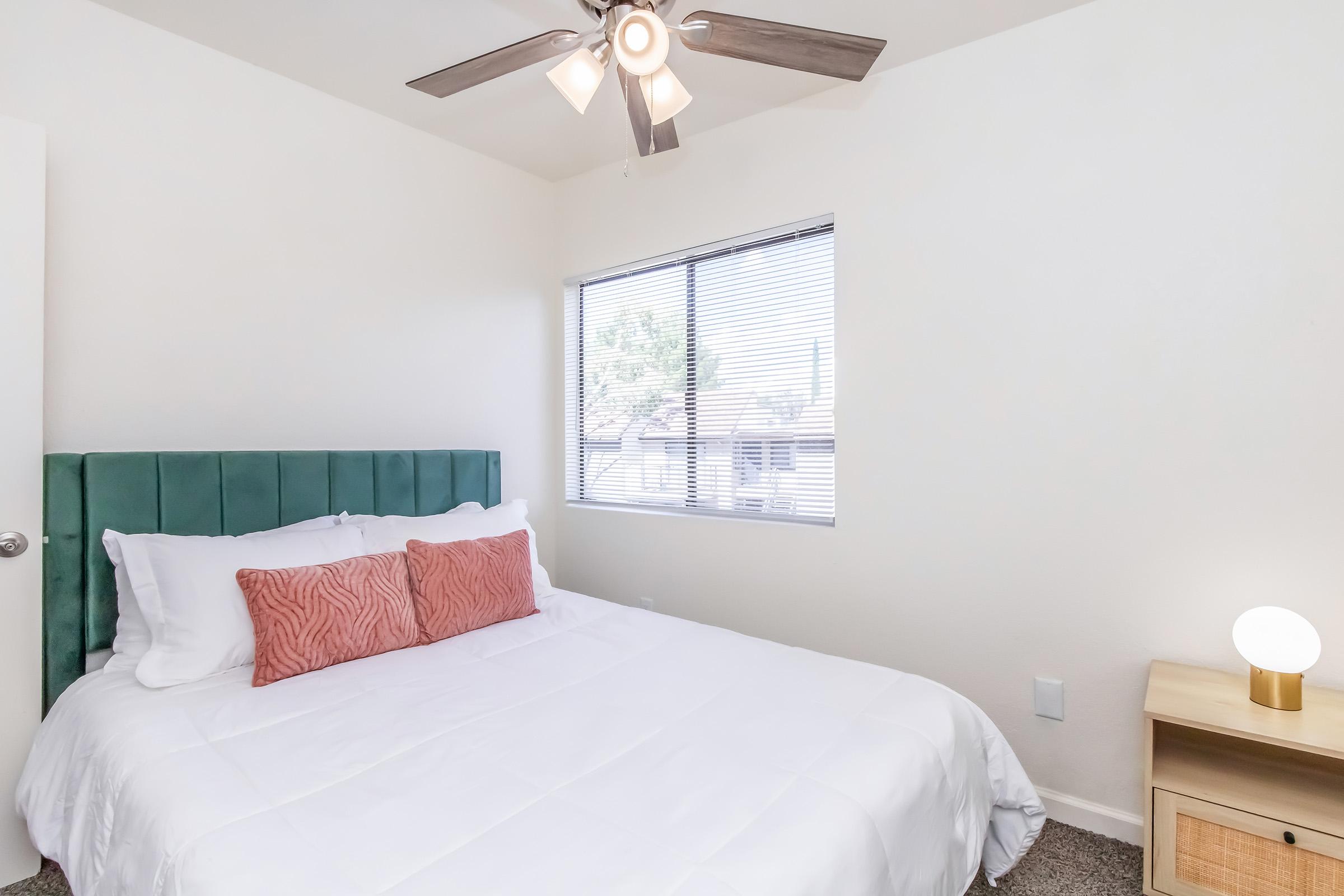 A well-lit bedroom featuring a comfortable queen-sized bed with white bedding and two decorative pillows. The room has a green upholstered headboard, a nightstand with a round lamp, and a window with blinds allowing natural light to enter. The walls are painted white, creating a bright and airy atmosphere.