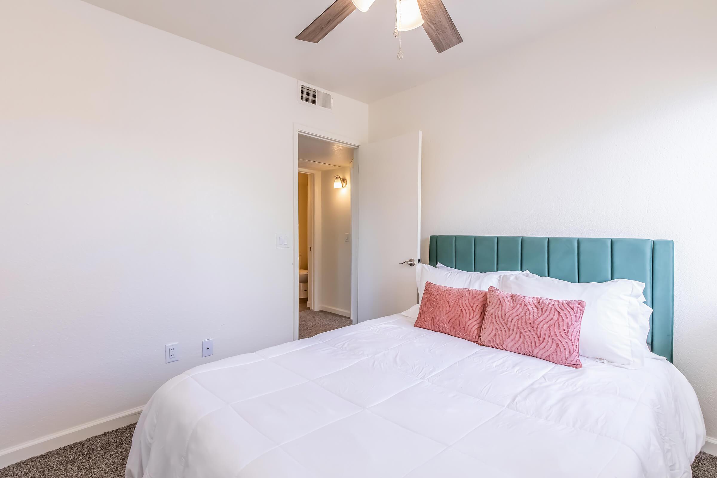 A well-lit bedroom featuring a white bed with a teal headboard and pink decorative pillows. The walls are painted white, and there is a ceiling fan overhead. A door leads to another room, with light visible from the doorway. The carpet is light-colored, adding to the room's cozy atmosphere.