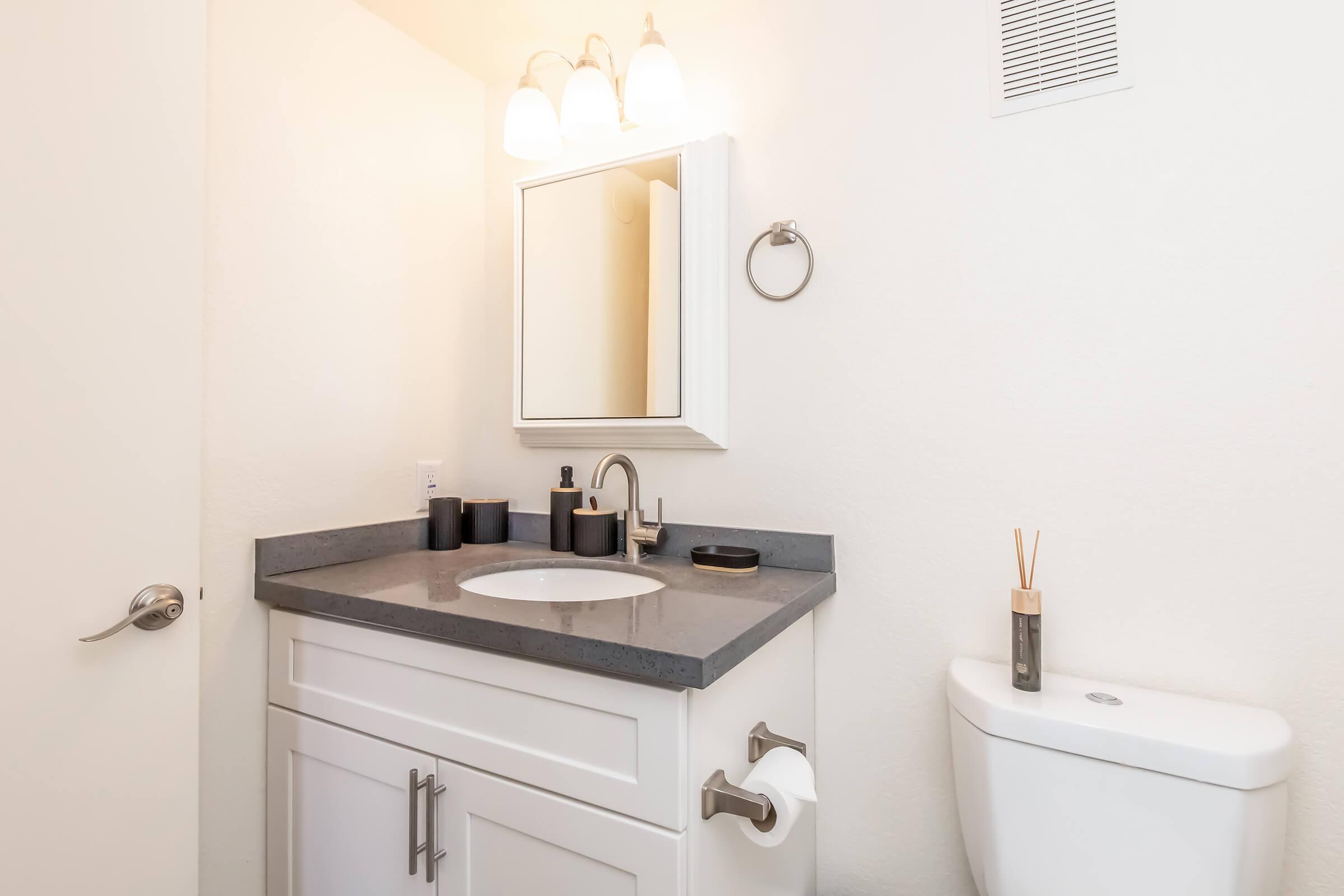 A bright and modern bathroom featuring a white vanity with a gray countertop, under-mounted sink, and a mirror. The space includes a stylish towel ring, decorative items, and a clean white toilet. Soft lighting from three fixtures adds warmth to the room.