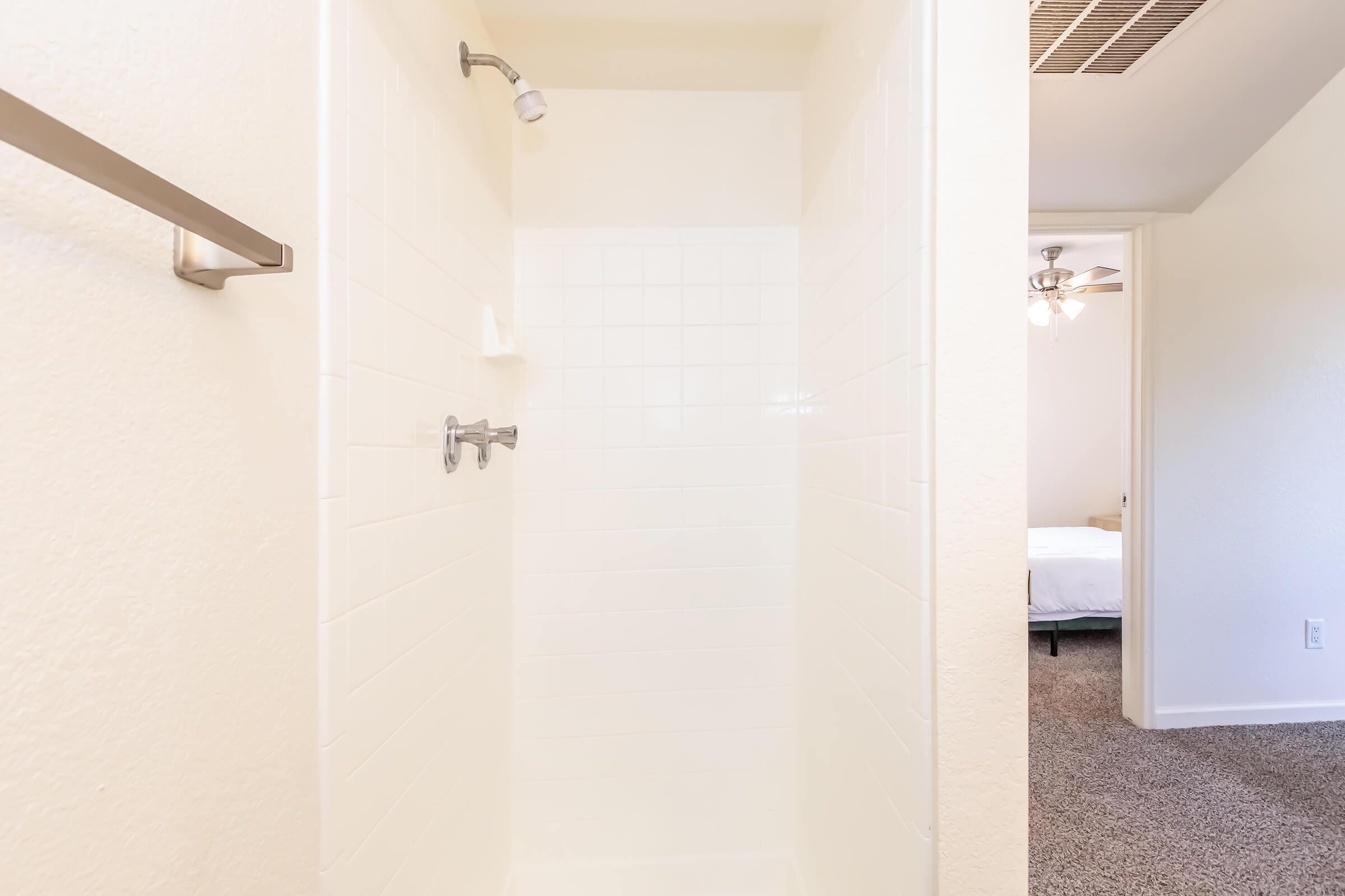 A clean, empty shower stall with white tiled walls and a chrome showerhead. A towel rack is mounted on the left wall. In the background, a glimpse of a bedroom with a ceiling fan and a white bed is visible. The space has a bright, airy feel with neutral-colored walls and carpeting.