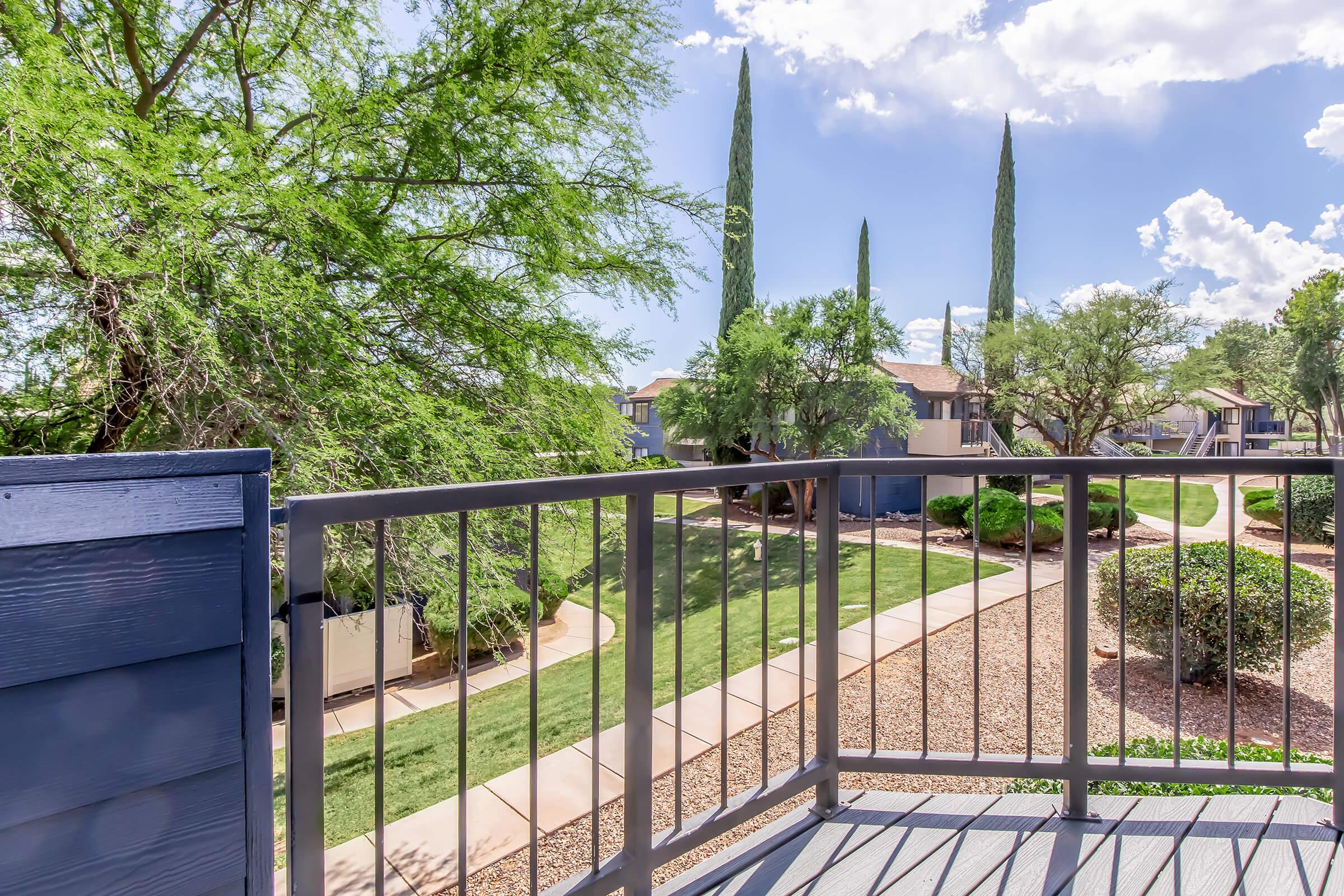 View from a balcony overlooking a landscaped area with walking paths, greenery, and tall conical trees under a bright blue sky with scattered clouds. The scene captures a peaceful residential setting, ideal for relaxation and enjoying the outdoors.