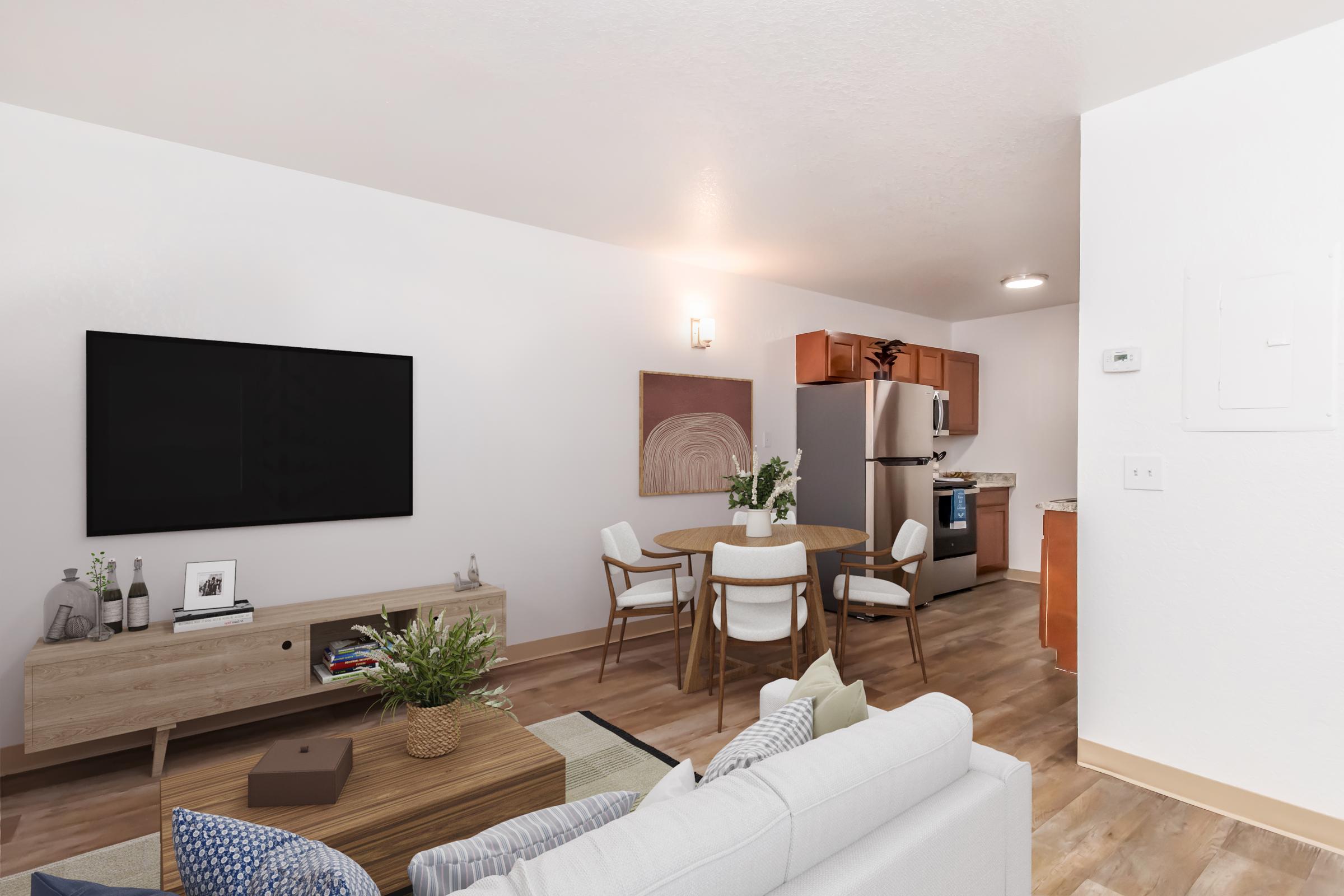 A modern living area featuring a light gray sofa, a wooden coffee table, and a large flat-screen TV mounted on the wall. In the background, there's a dining area with a round table and white chairs, alongside a kitchen with wooden cabinetry and stainless steel appliances. Natural light fills the space.
