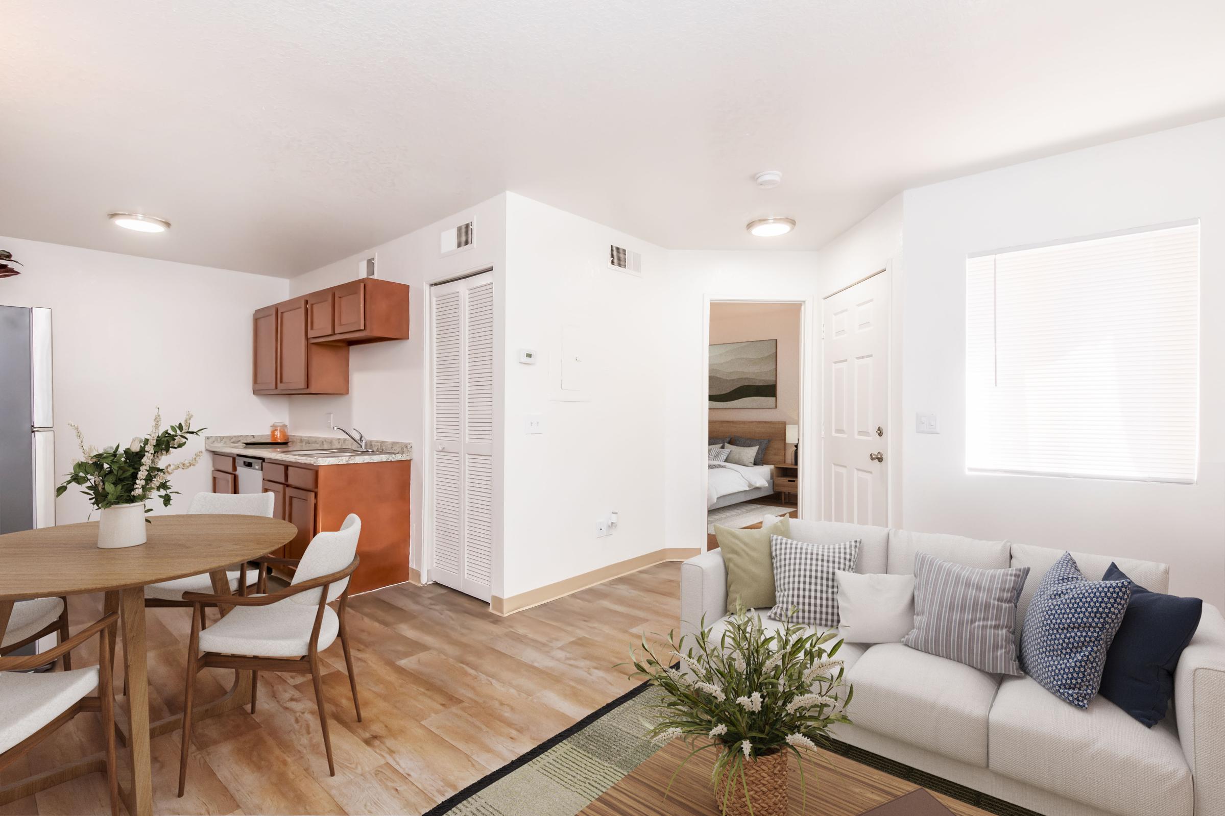 A modern, well-lit living space featuring a cozy sofa adorned with decorative pillows, a circular dining table surrounded by chairs, and an open kitchen with wooden cabinets. A doorway leads to a bedroom, and large windows allow natural light to fill the room. The decor is simple and inviting, creating a warm atmosphere.