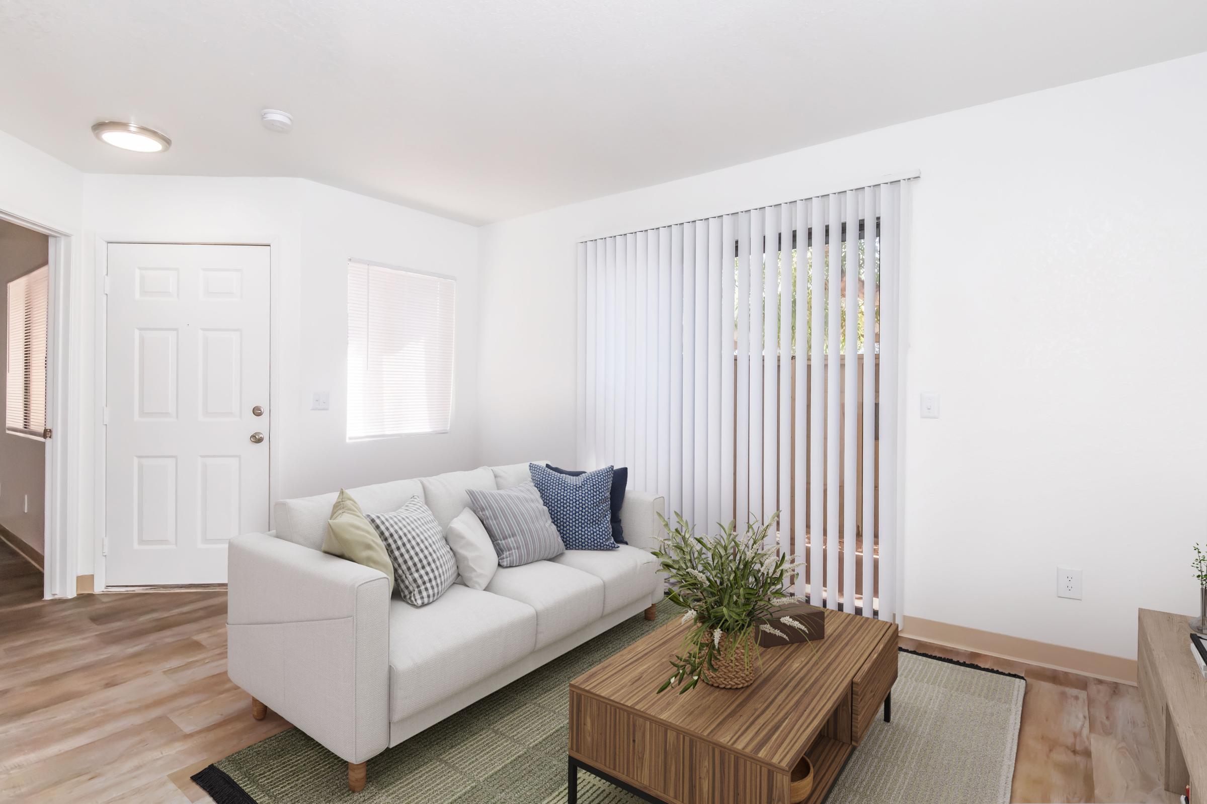 A cozy living room featuring a light-colored sofa with decorative pillows, a wooden coffee table, and a green plant. Large windows with vertical blinds allow natural light to fill the space. The floor has wooden planks, and the walls are painted in a soft white, giving a modern and inviting feel.