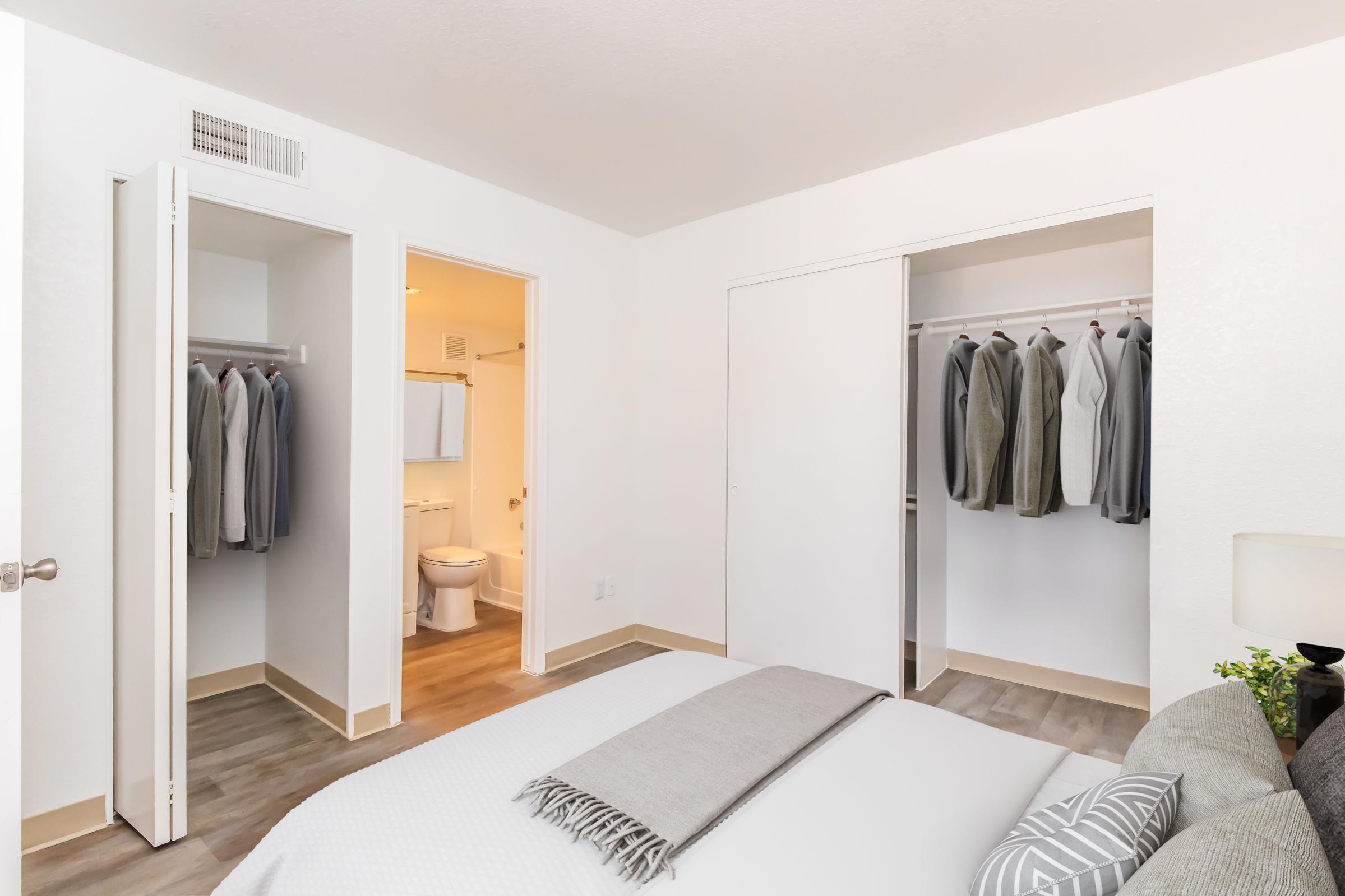 A bright, modern bedroom featuring a neatly made bed with a light gray blanket. On the left, a closet with hanging clothes is visible, and a bathroom door is open on the opposite side. The room has a minimalist design, with light walls and wooden flooring, complemented by a small lamp on a bedside table.