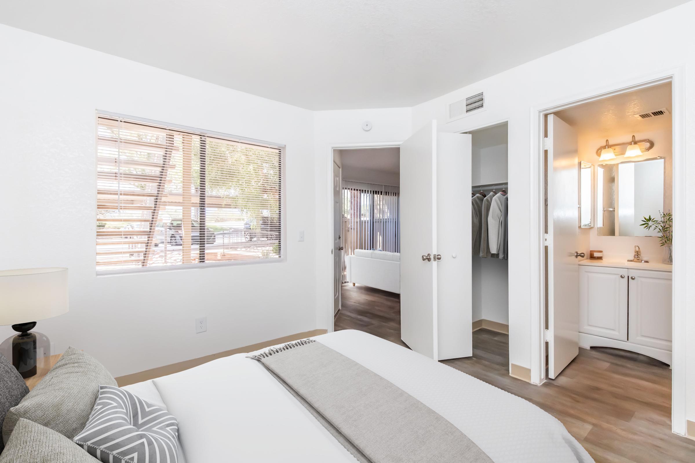 A bright, modern bedroom featuring a neatly made bed with decorative pillows, a window with blinds, and an open closet with hanging clothes. The room includes a doorway leading to a bathroom and another area, with light wooden flooring and neutral-colored walls.