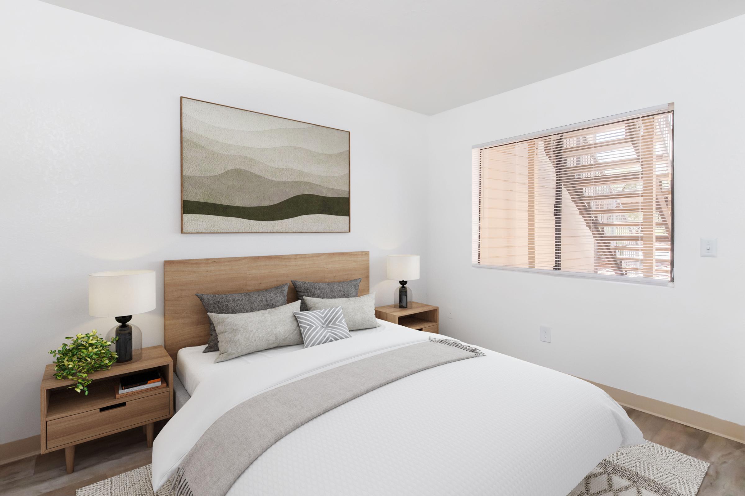 A modern bedroom featuring a wooden bed with a light gray blanket and decorative pillows, two bedside tables with lamps, and a green plant. A large artwork with abstract shapes hangs on the wall, and a window covered with blinds allows natural light to enter the room.