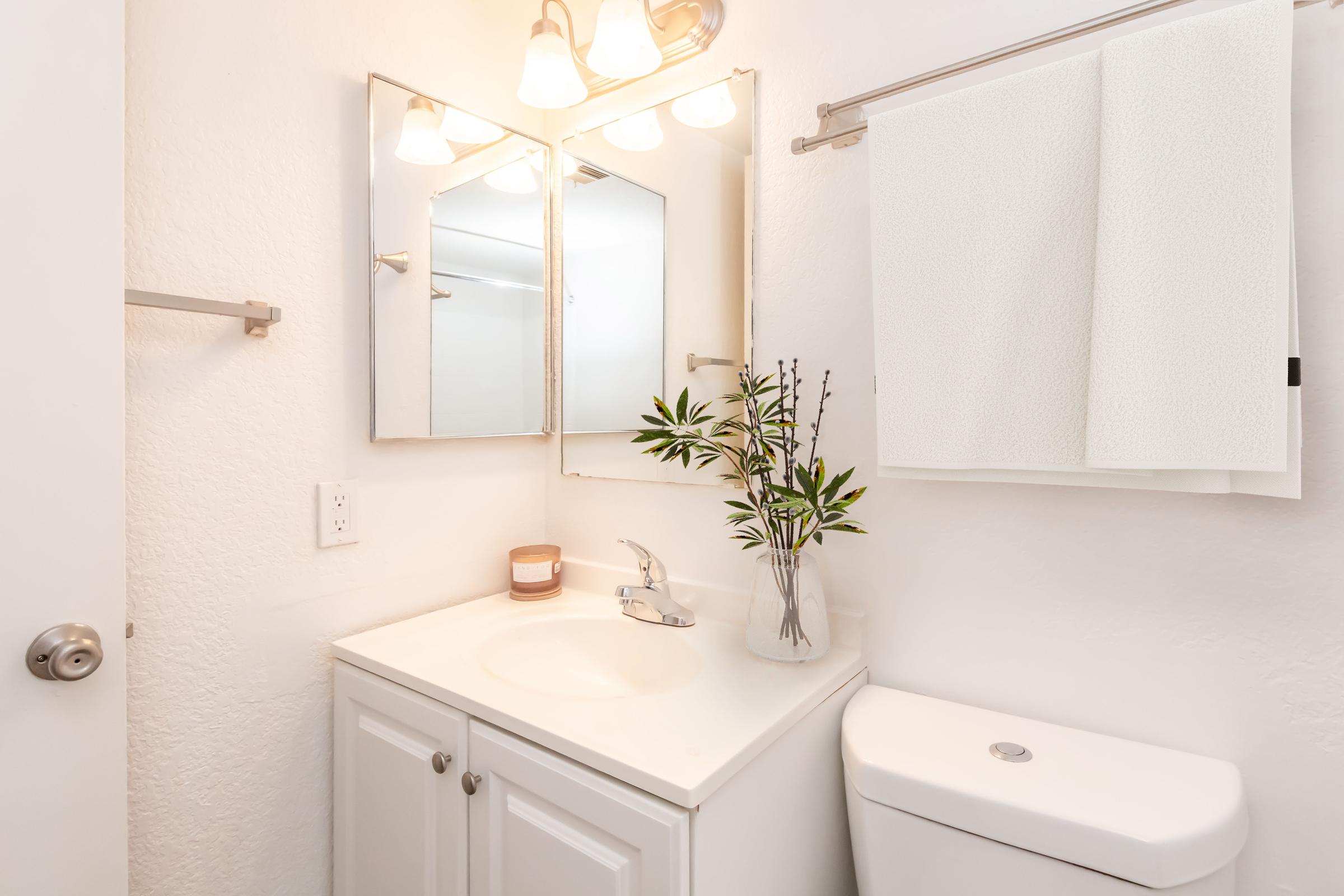 A clean, modern bathroom featuring a white vanity with a sink, a wall-mounted mirror with three light fixtures above, a white towel hanging on a silver rod, and a small plant in a vase. A toilet is positioned beside the vanity against a light-colored wall, creating a bright and airy atmosphere.