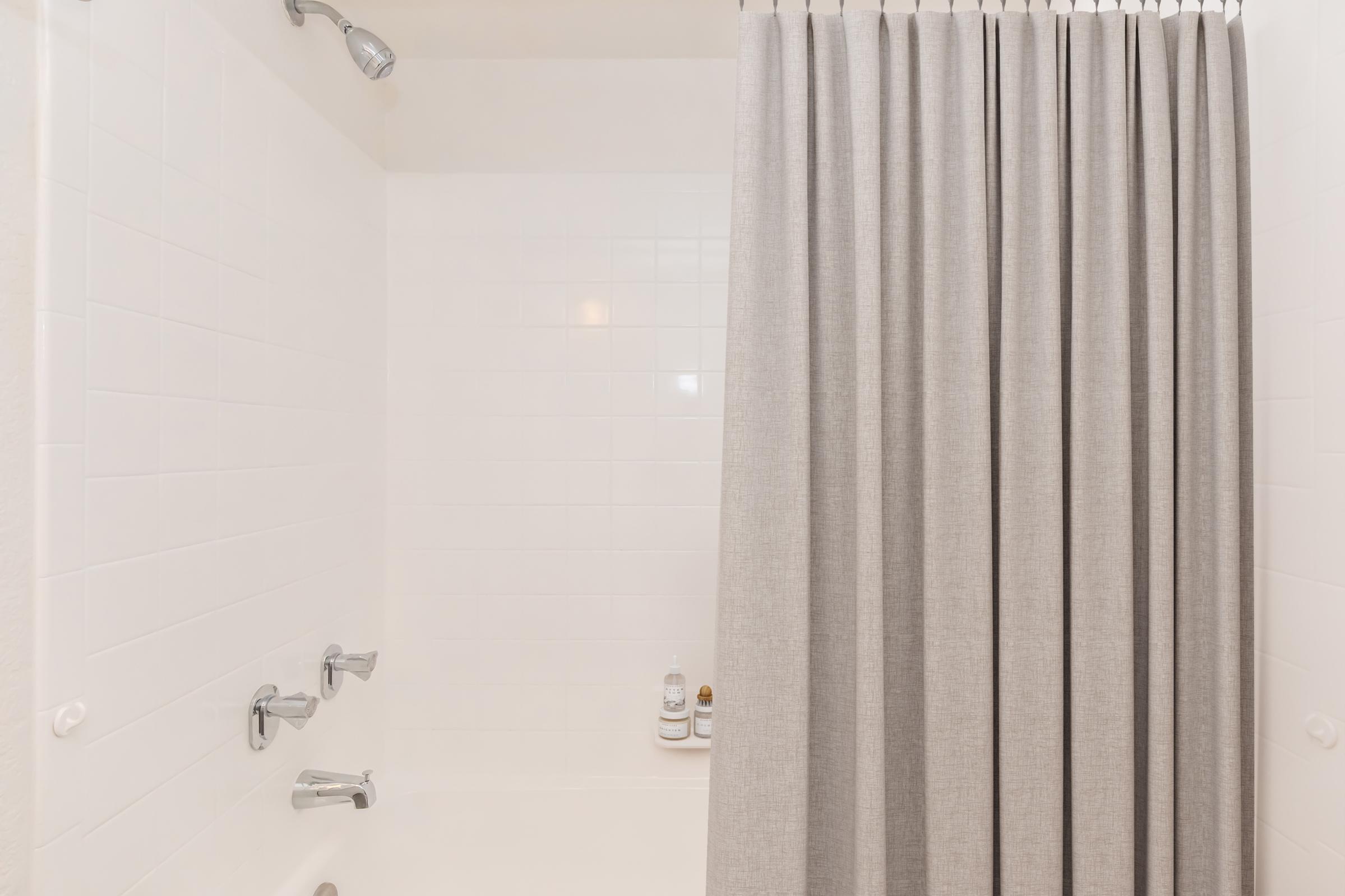 A clean bathroom with a shower area featuring a simple gray curtain. The walls are tiled in white, and there are chrome fixtures for the shower and faucet. A couple of shampoo bottles are placed on the edge of the bathtub. The overall aesthetic is minimalist and tidy.