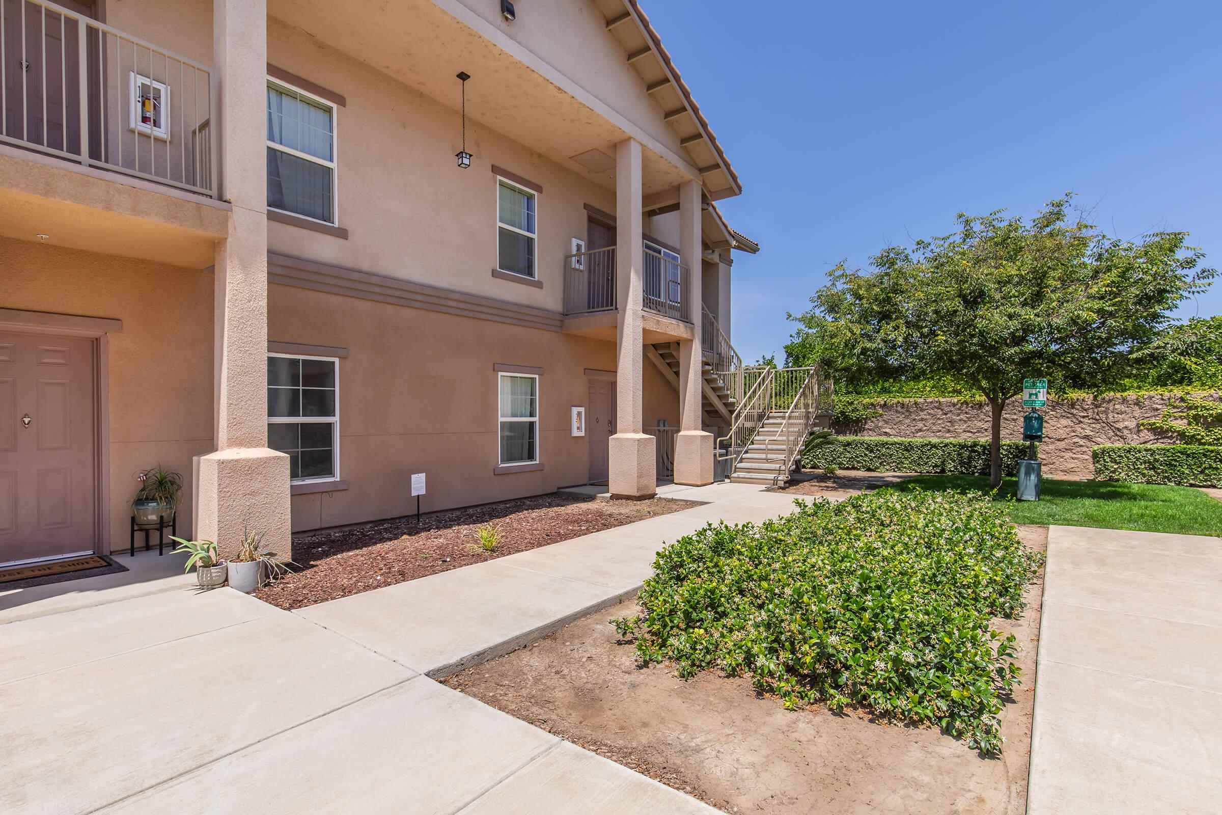 Exterior view of a multi-unit apartment building featuring a beige facade, a set of stairs leading to the upper level, and a landscaped area with small shrubs. The entrance has a door with a simple design, and there are clear skies in the background, indicating a sunny day.