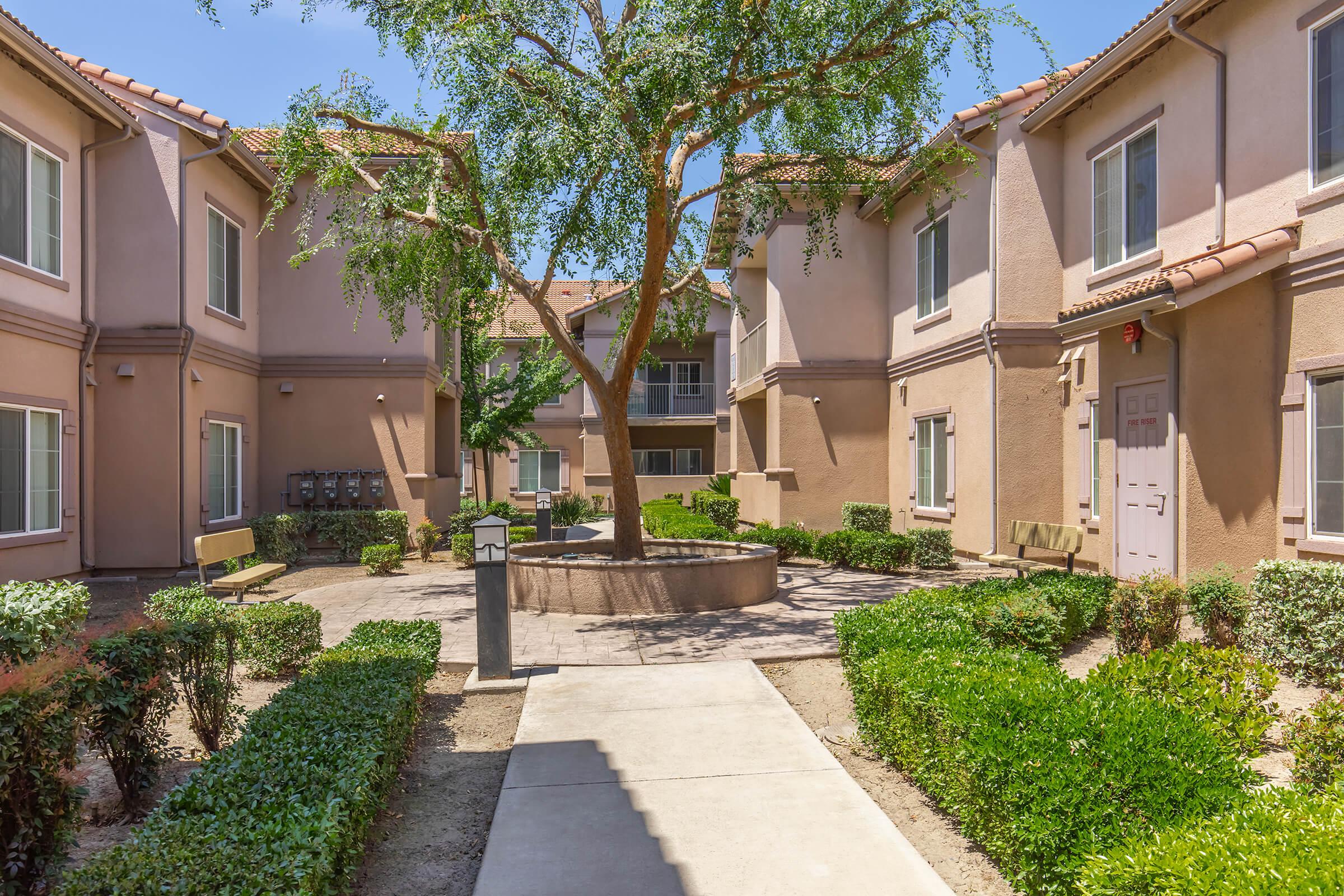 A landscaped courtyard of an apartment complex featuring a central tree, surrounded by bushes and benches. Pathways lead through the green space, connecting several buildings with beige exteriors and windows. The scene is well-lit under a clear blue sky.