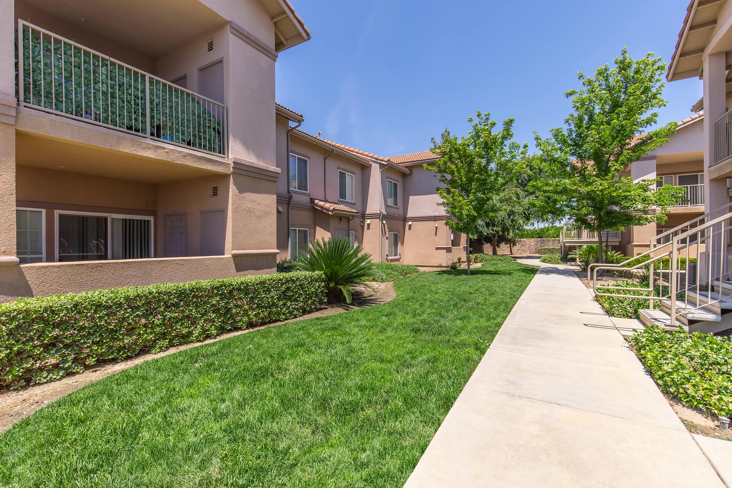 A well-maintained residential complex featuring two-story buildings with balconies, surrounded by lush green lawns and landscaped bushes. A wide concrete walkway runs between the buildings, leading towards a sunny area with trees, creating a pleasant neighborhood atmosphere.