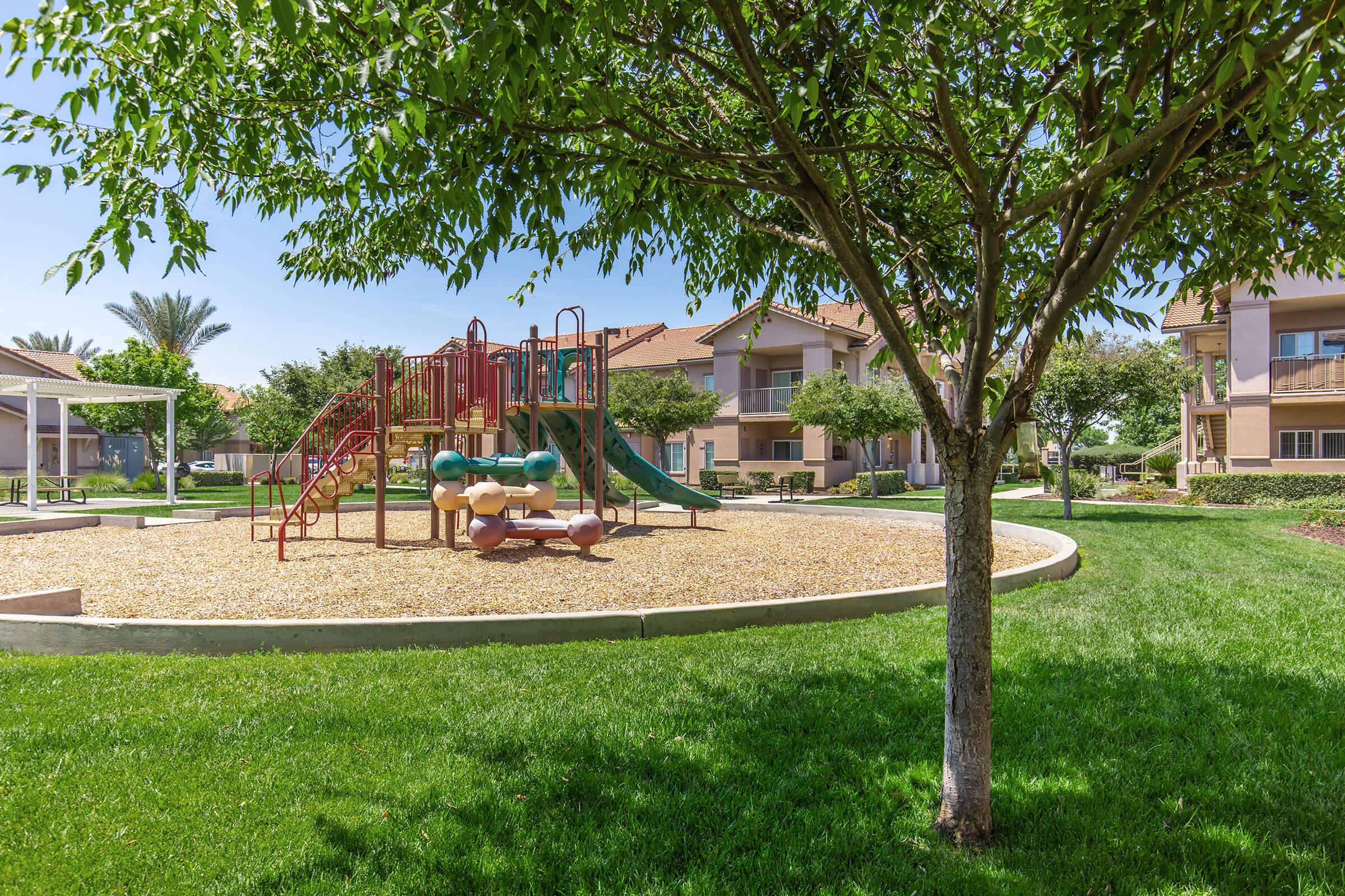 A bright playground featuring a red and green climbing structure with slides, surrounded by soft grass and shaded by trees. Nearby, residential buildings with balconies can be seen under a clear blue sky, creating a welcoming outdoor space for children and families.