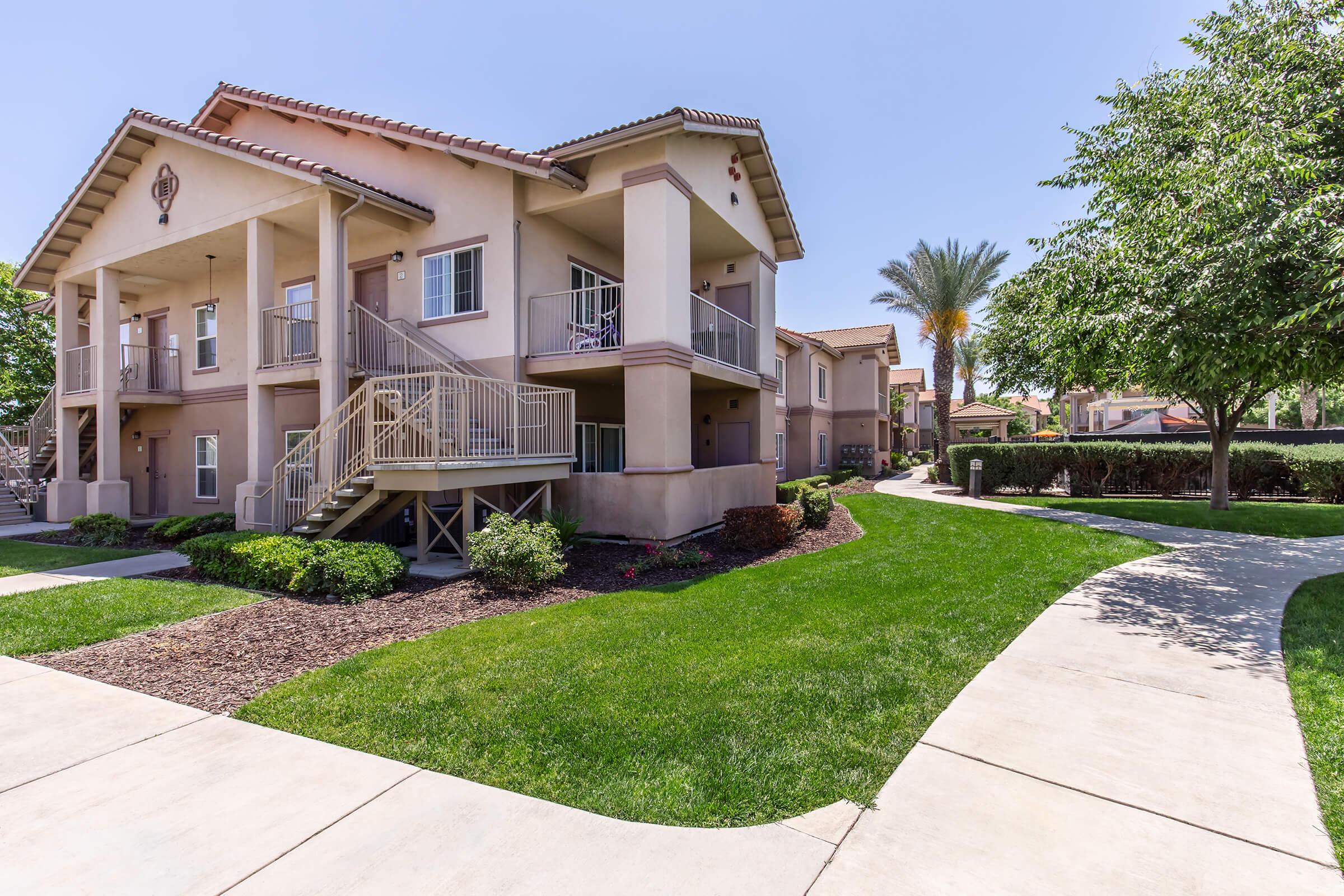 Two-story residential apartment building with outdoor staircases and balconies. Lush green lawn and well-maintained landscaping surround the building. A paved walkway leads through the grass, with palm trees and shrubs in the background under a clear blue sky.