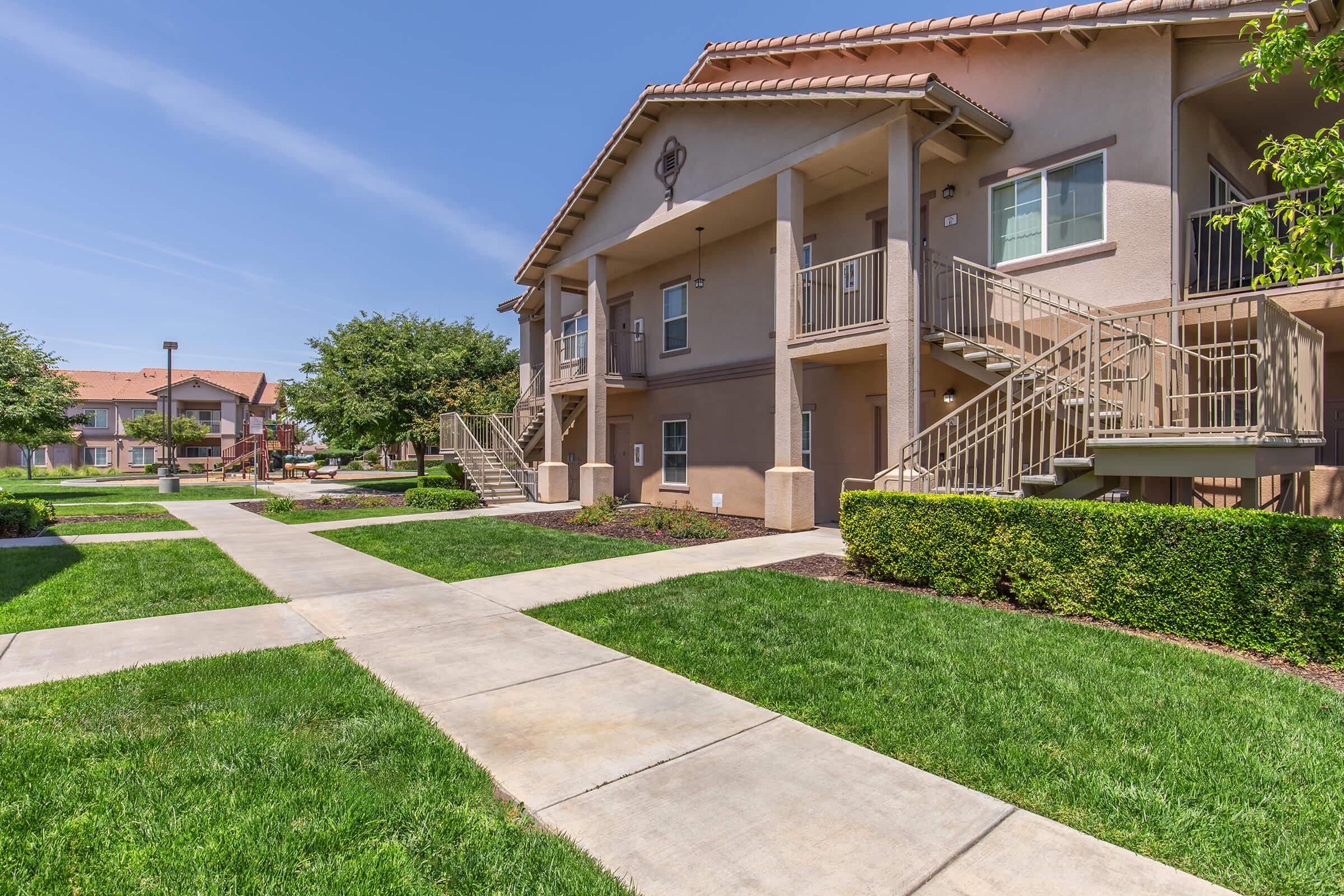 Exterior view of a modern multi-unit apartment complex. The image features two attached buildings with patios, surrounded by manicured lawns and walkways. Clear blue sky overhead and well-maintained landscaping enhance the inviting atmosphere of the residential area.