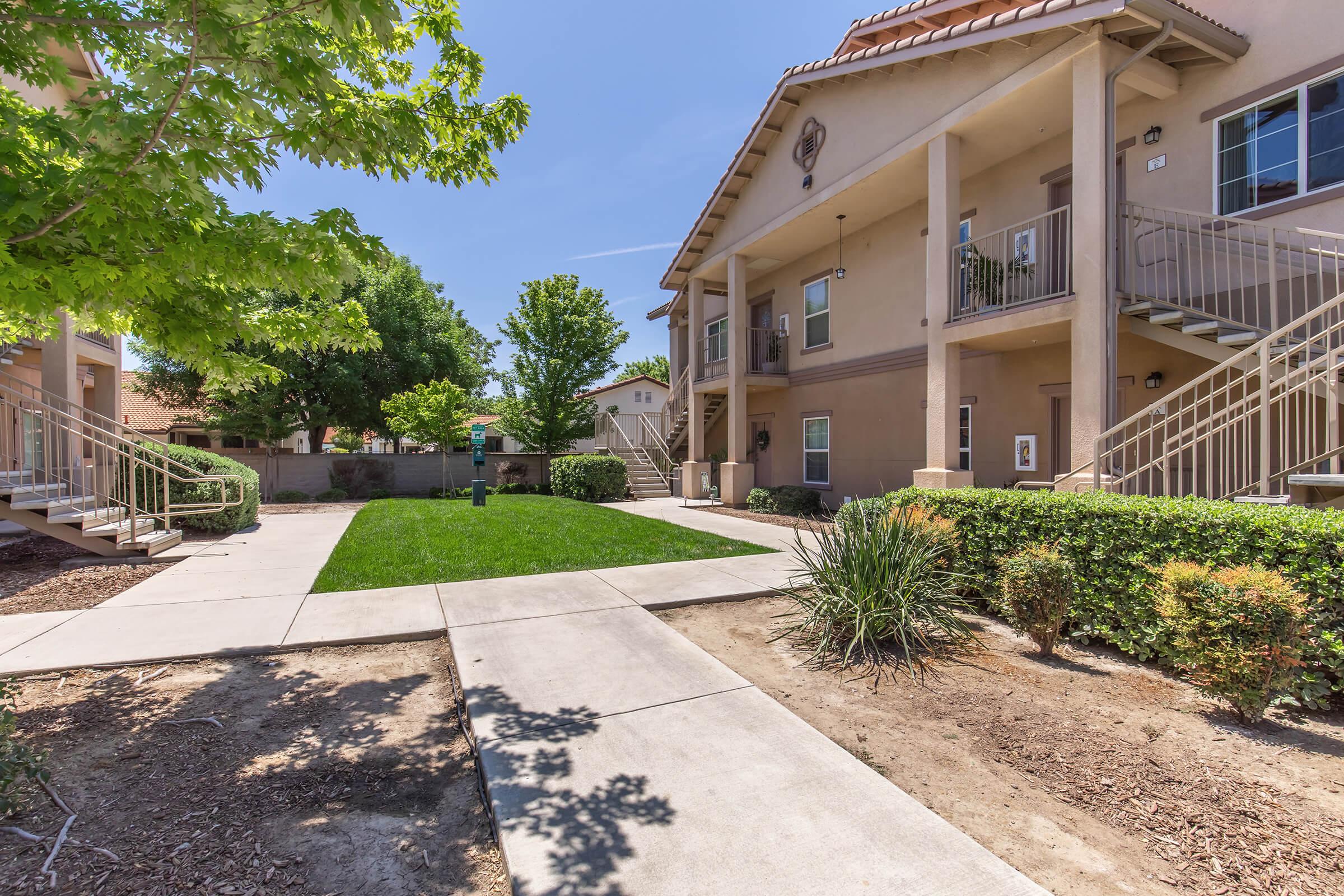 A view of a sunny apartment complex featuring two-story buildings, green lawns, and landscaped pathways. Trees provide shade, and there are staircases leading to the upper level of the apartments. The scene is well-maintained and inviting, with a clear blue sky in the background.