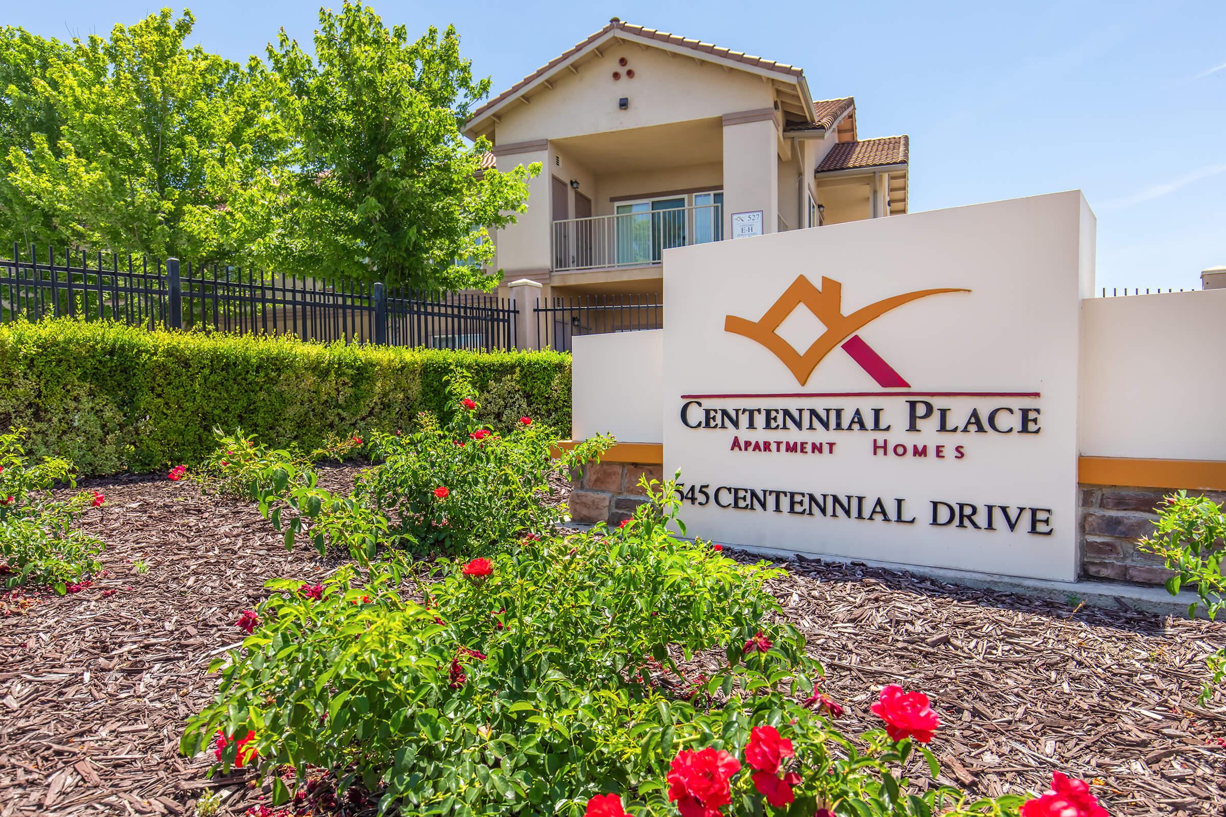 A sign for Centennial Place Apartment Homes located at 645 Centennial Drive, surrounded by landscaped greenery and flowering bushes. A building is visible in the background under a clear blue sky.