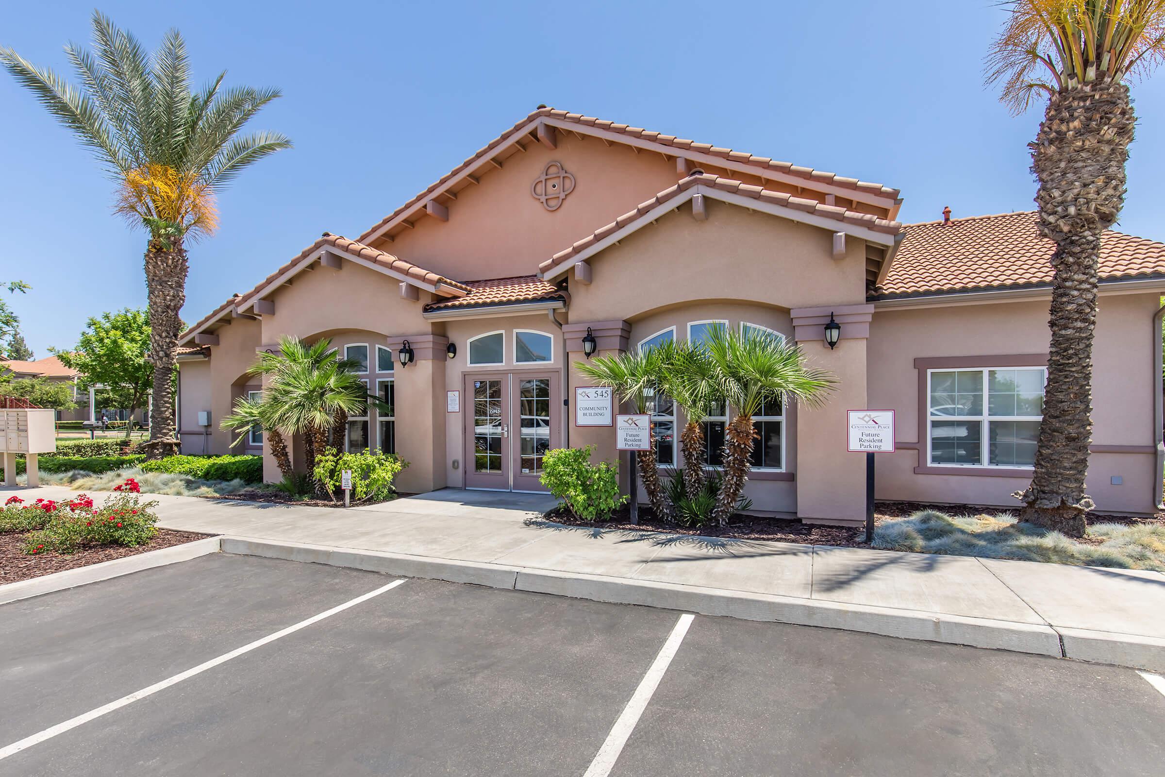 A single-story building with a Mediterranean-style architecture featuring a tiled roof, large windows, and palm trees in front. The entrance is framed by two entrance doors, and the surrounding area includes well-maintained landscaping with flowers and a paved parking lot. Bright, clear blue sky overhead.