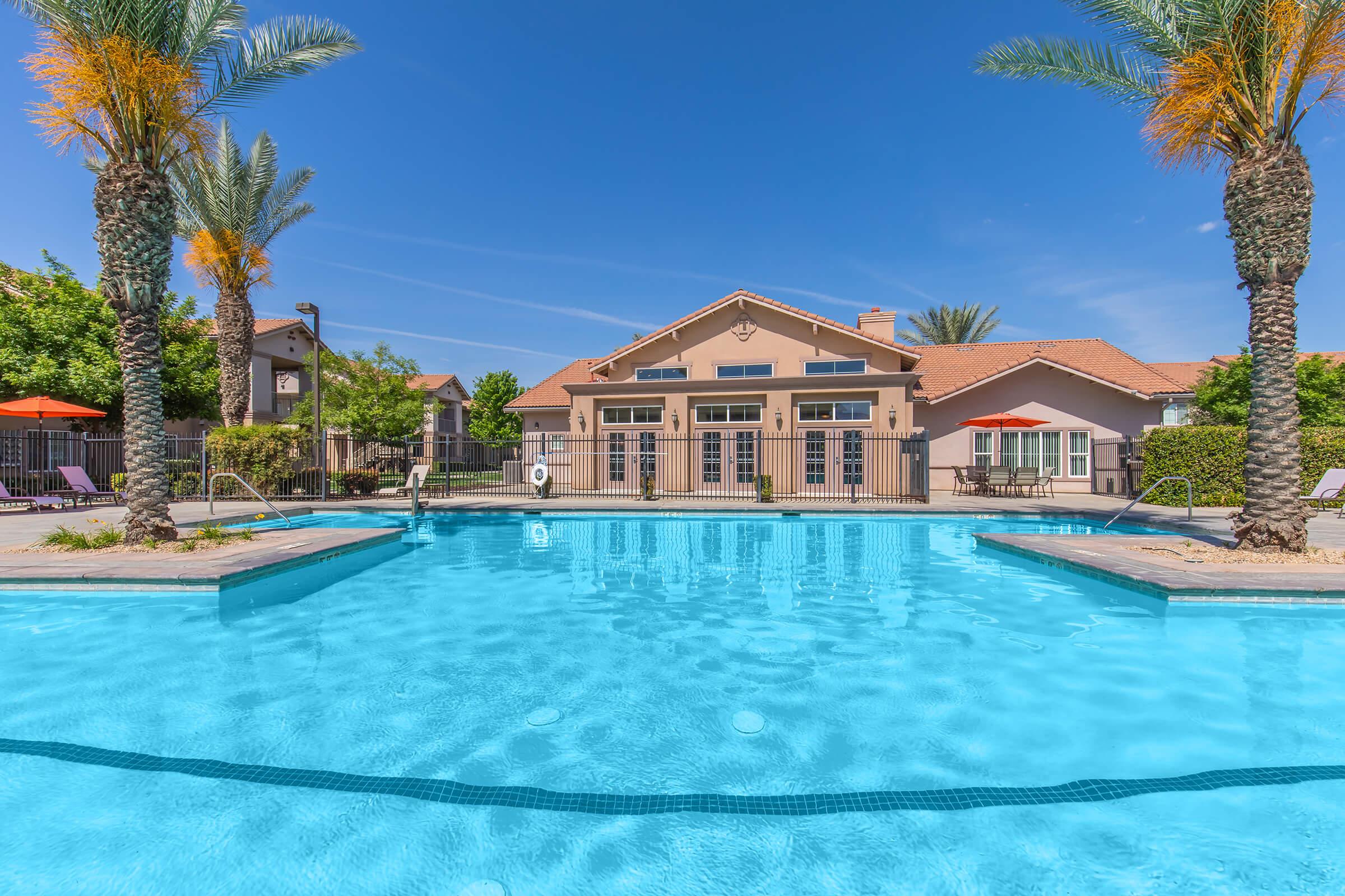 A clear blue swimming pool surrounded by palm trees and lounge chairs, with a well-maintained building in the background. The sky is bright and sunny, creating a relaxing outdoor atmosphere.