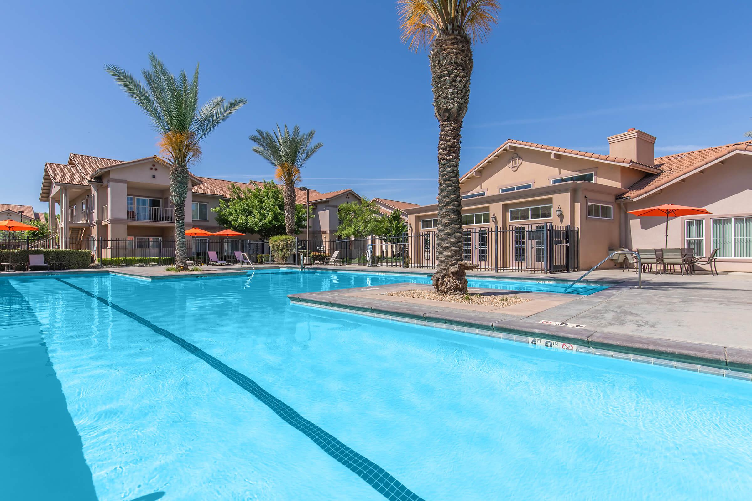 A bright outdoor swimming pool surrounded by palm trees and lounge chairs. In the background, a residential building with orange umbrellas can be seen. The clear blue water reflects the sunny sky, creating a relaxing atmosphere.
