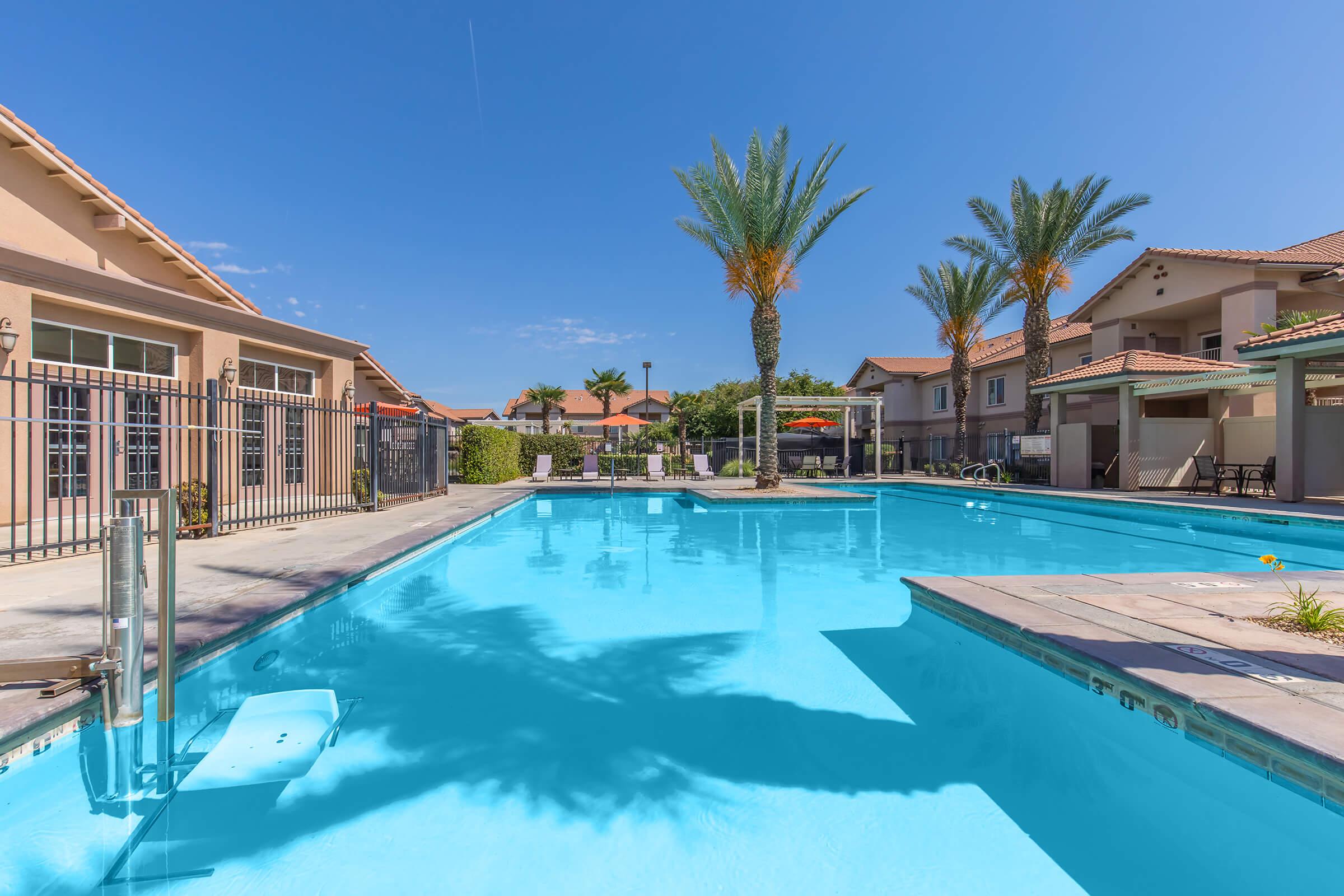 A tranquil swimming pool surrounded by palm trees and residential buildings. The clear blue water reflects the bright sky, and lounge chairs are positioned around the pool area, creating a relaxing outdoor space. The area is well-maintained with lush greenery in the background.