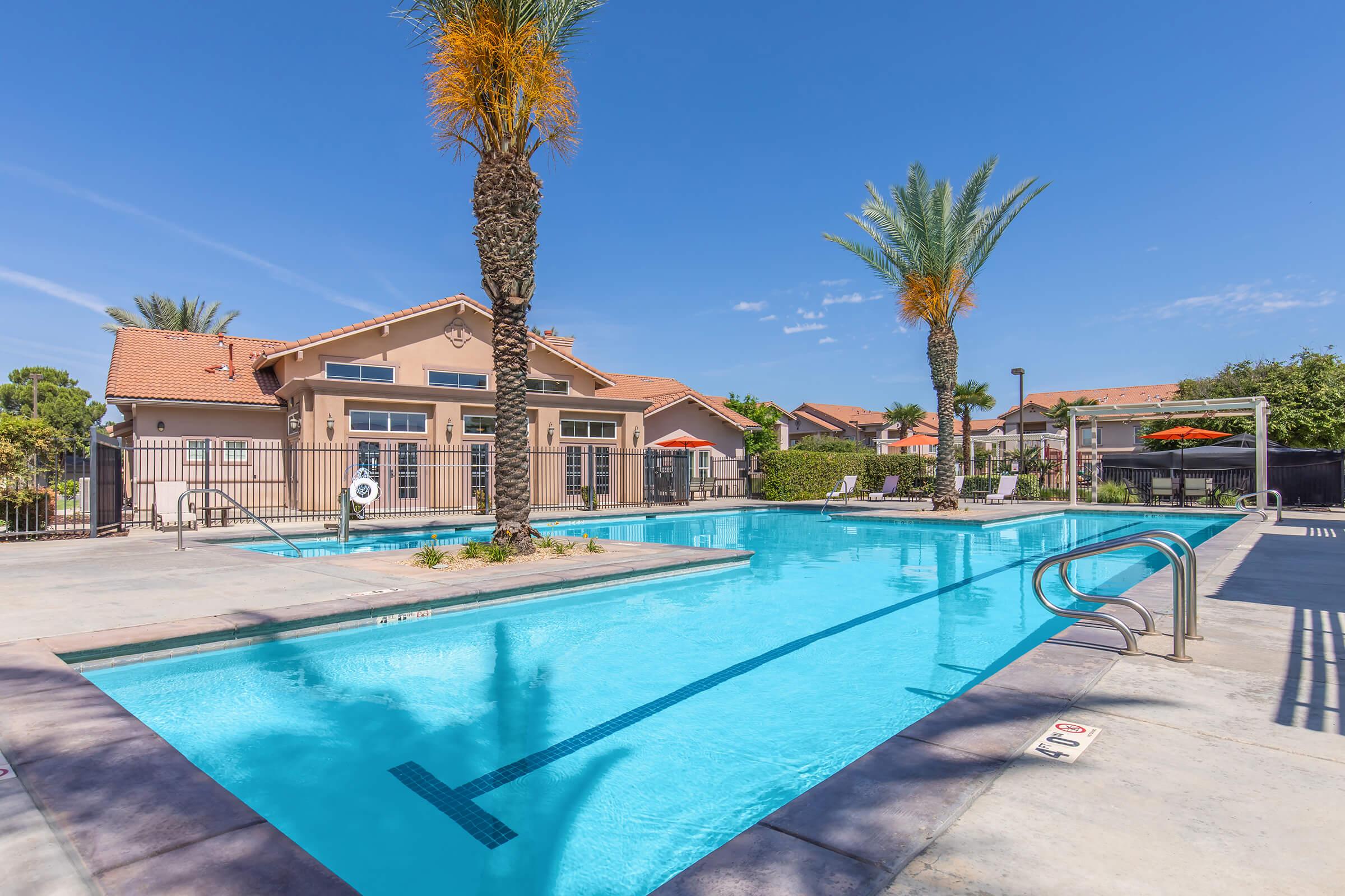 A clear blue swimming pool surrounded by palm trees and lounge chairs, with a gated area and a residential building in the background under a sunny sky. The pool features steps leading in and a shallow area.