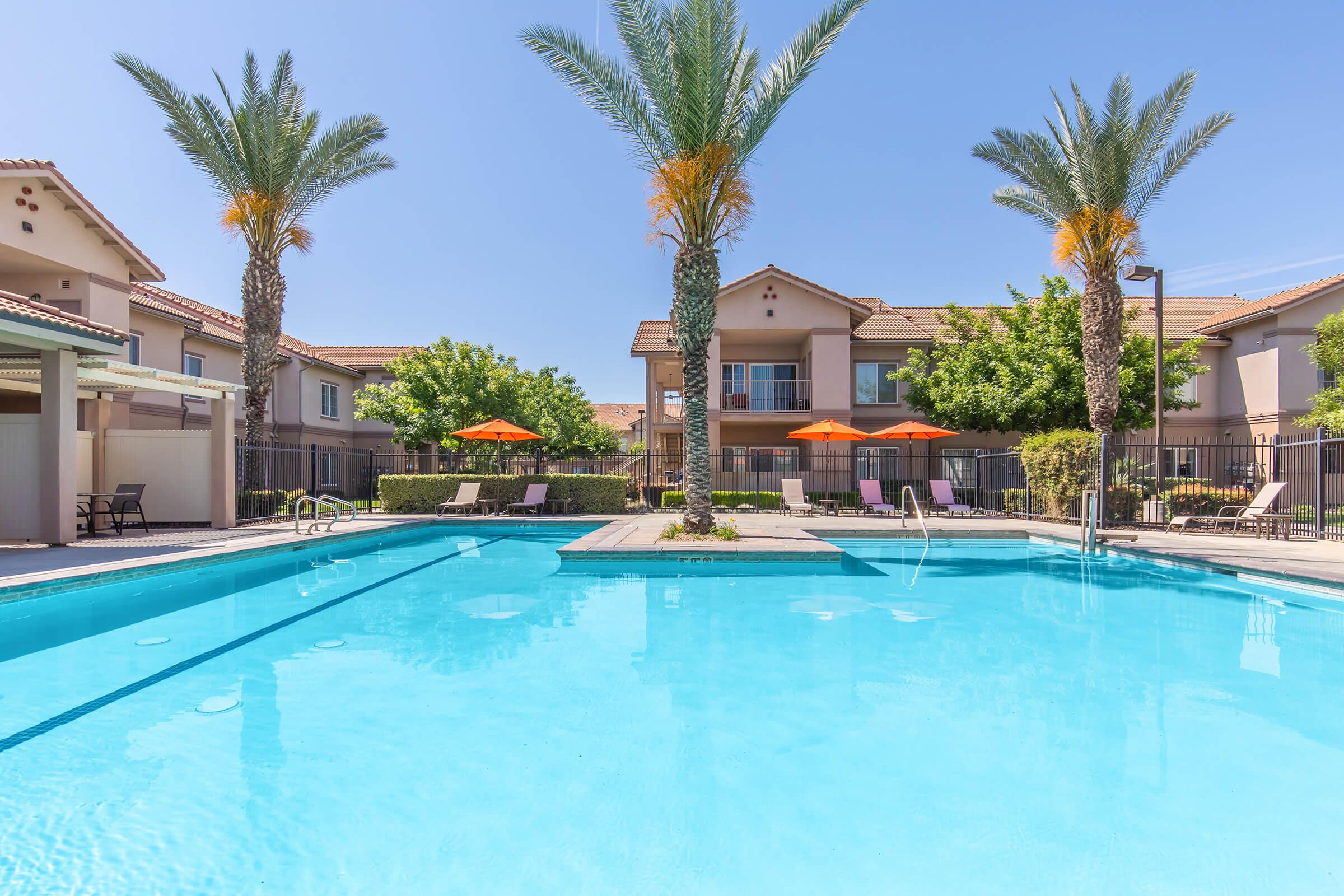 A bright blue swimming pool surrounded by palm trees and lounge chairs. In the background, there are two-story buildings with orange umbrellas providing shade. The clear sky adds to the sunny, inviting atmosphere of the pool area.