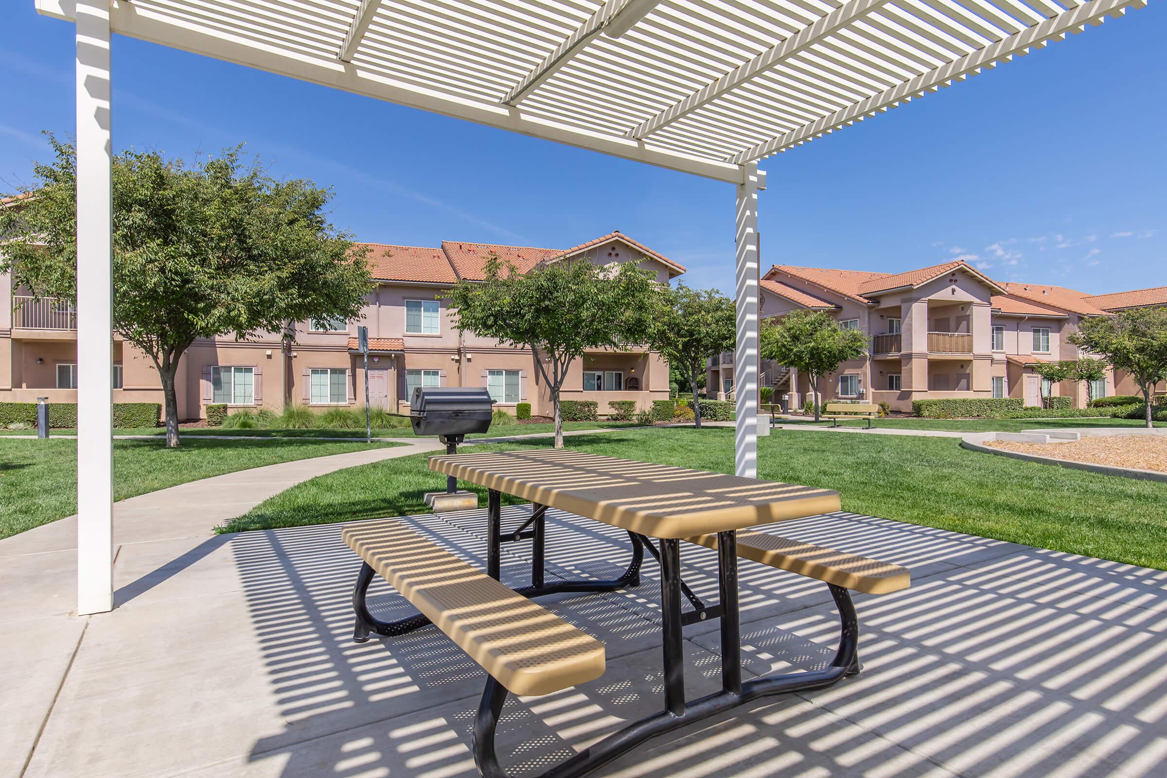 A picnic table under a white pergola in a landscaped area with green grass. Apartment buildings with orange roofs are in the background under a clear blue sky, and there are trees providing shade nearby. A barbecue grill can also be seen in the vicinity.