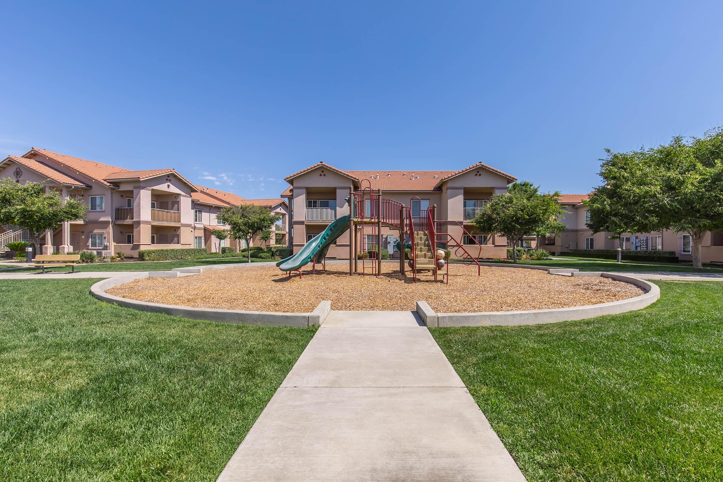 A playground featuring a slide and climbing structure is centered in a grassy area surrounded by several apartment buildings with orange roofs under a clear blue sky. A concrete walkway leads to the playground, providing access for residents and visitors.