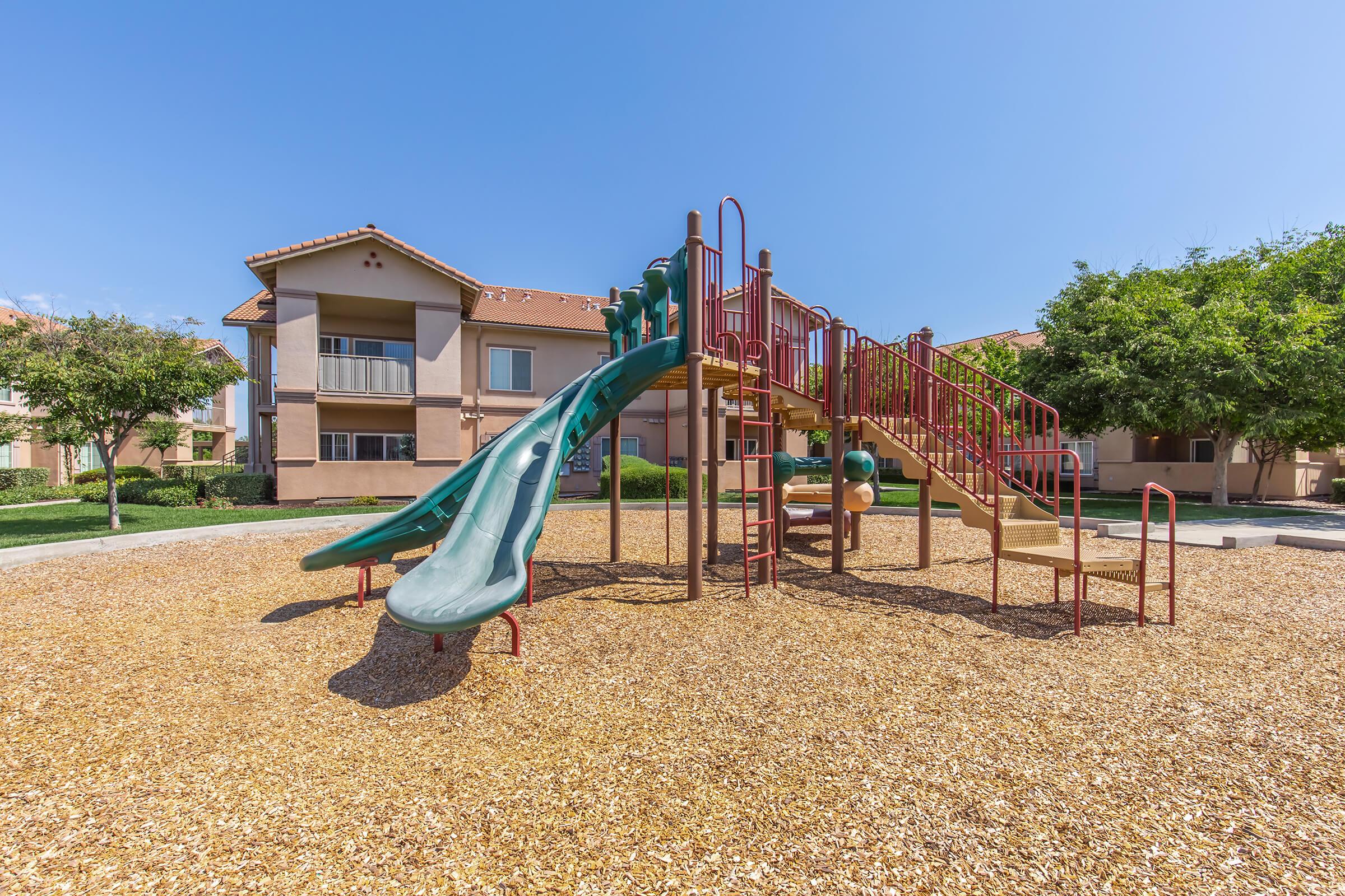 A playground featuring a green slide and climbing structure, surrounded by wood chip flooring. In the background, there are residential buildings and several trees, under a clear blue sky. The area is well-maintained and inviting for children to play.