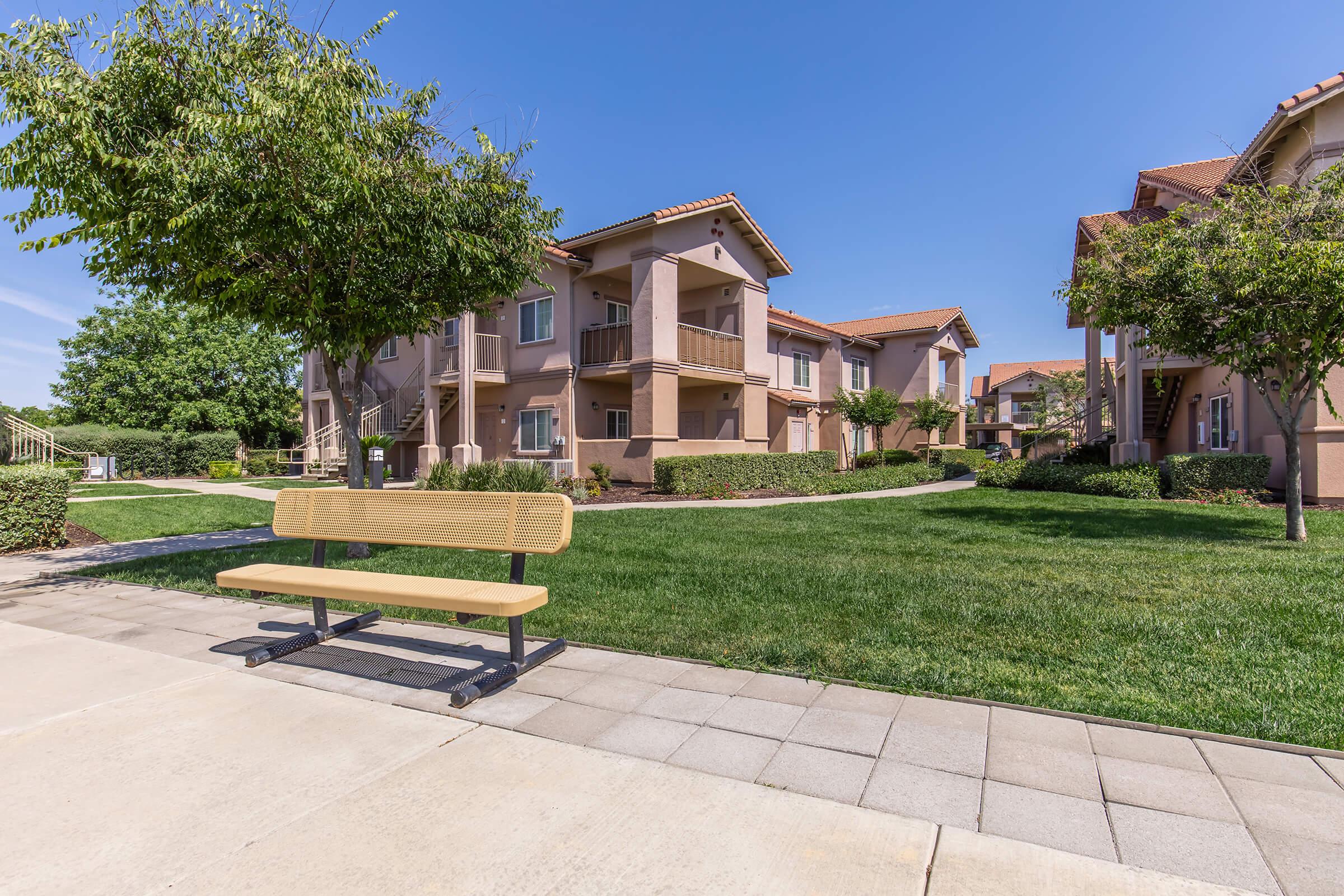A sunny outdoor view of an apartment complex featuring landscaped green lawns and trees. In the foreground, there is a wooden bench on a paved path, while the buildings are shown in soft pastel colors with balconies and a clear blue sky above.
