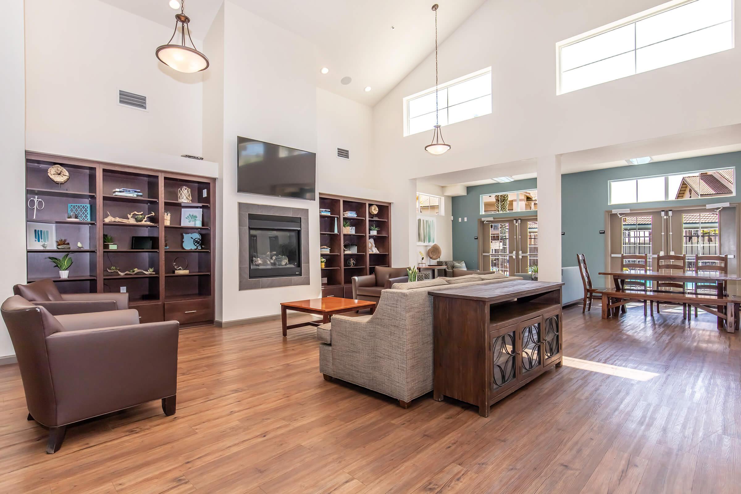 A spacious, well-lit living area with high ceilings, featuring a stylish fireplace, built-in bookshelves, and large windows. Comfortable seating includes a brown armchair and modern sofa. A wooden coffee table is center stage, and a dining area with chairs is visible in the background, enhancing the inviting ambiance.